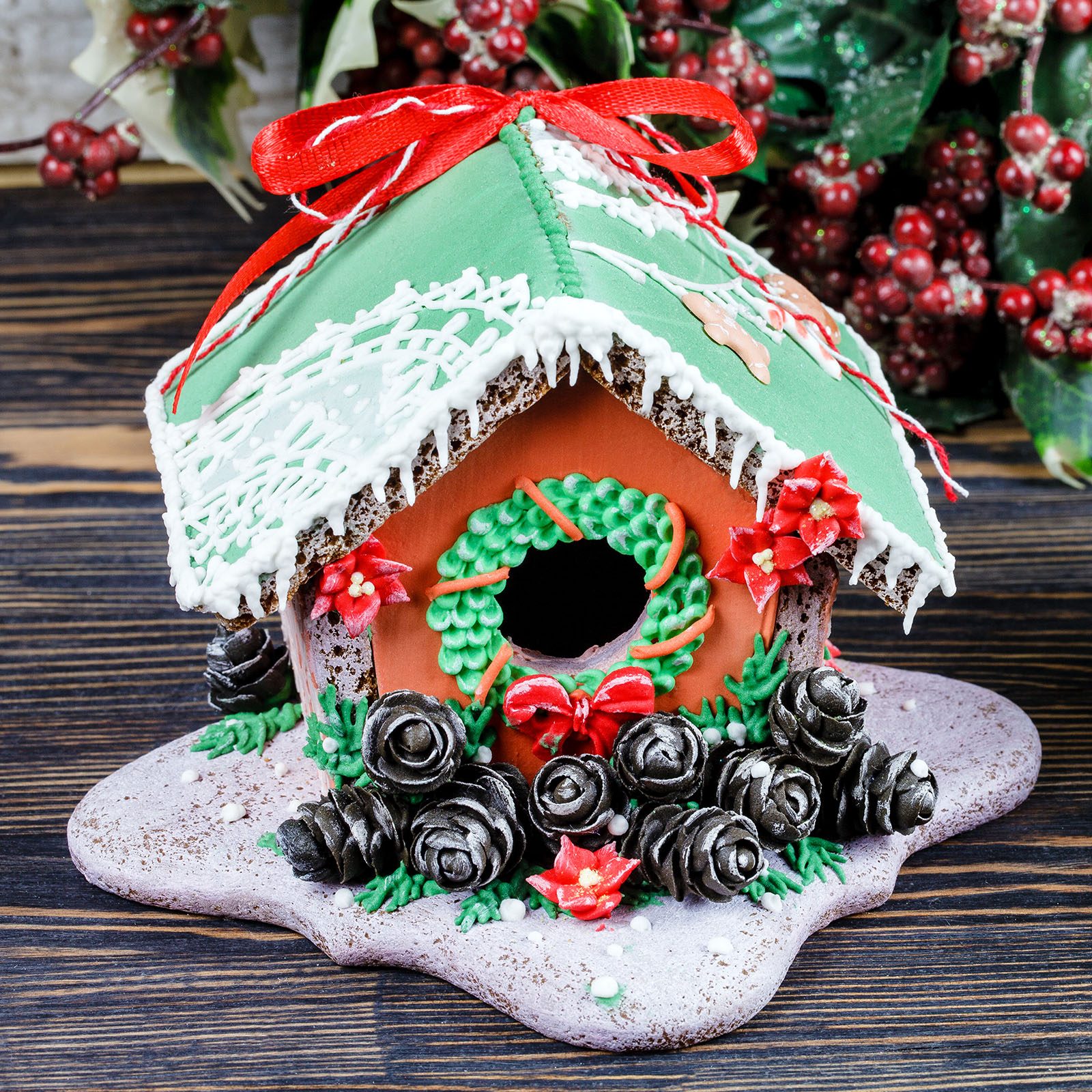 Gingerbread house decorated with icing and candy wreath, surrounded by chocolate pine cones, set on a wooden table with festive berries in the background.