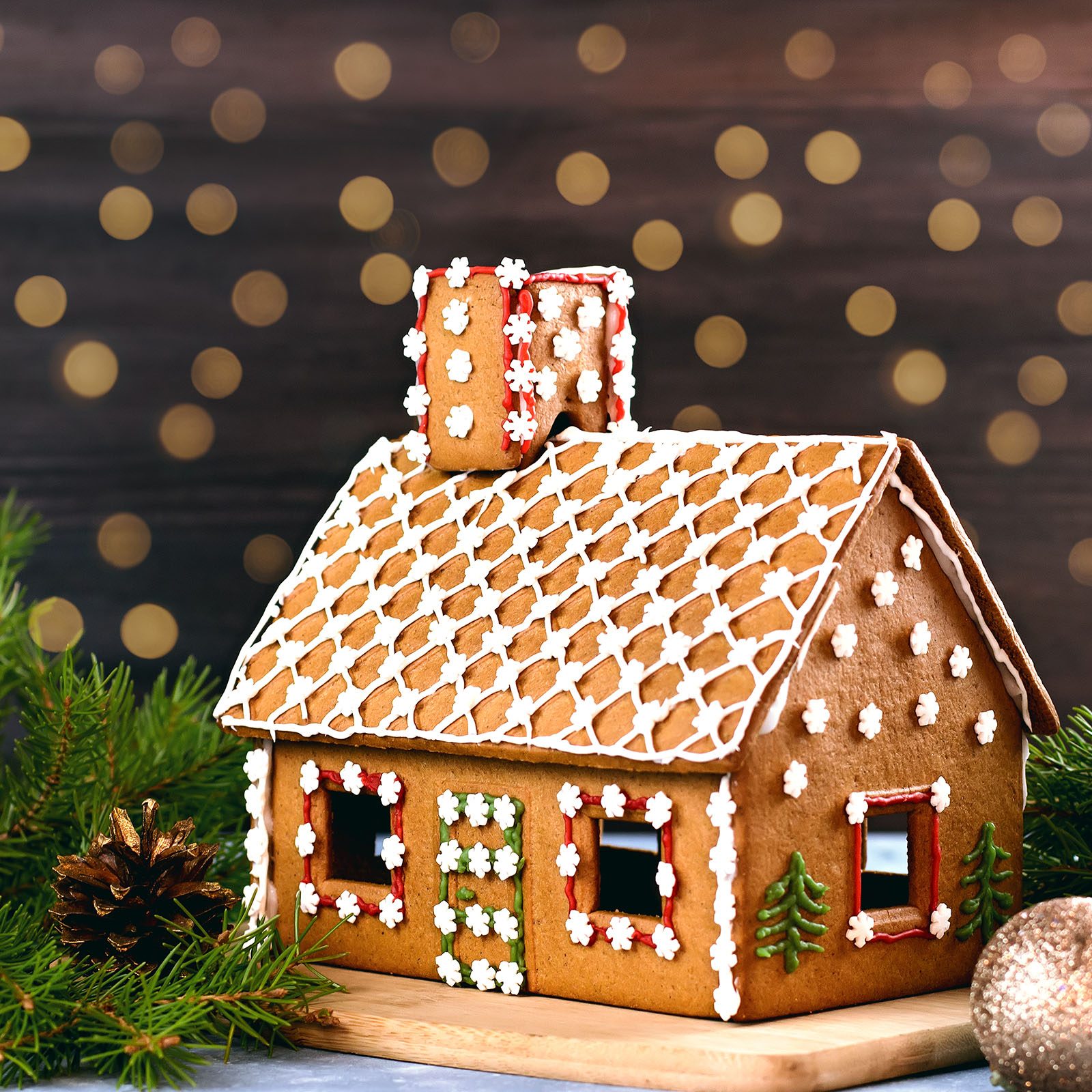 Gingerbread house adorned with white icing patterns sits on a wooden board, surrounded by festive pine branches and a glittering background.