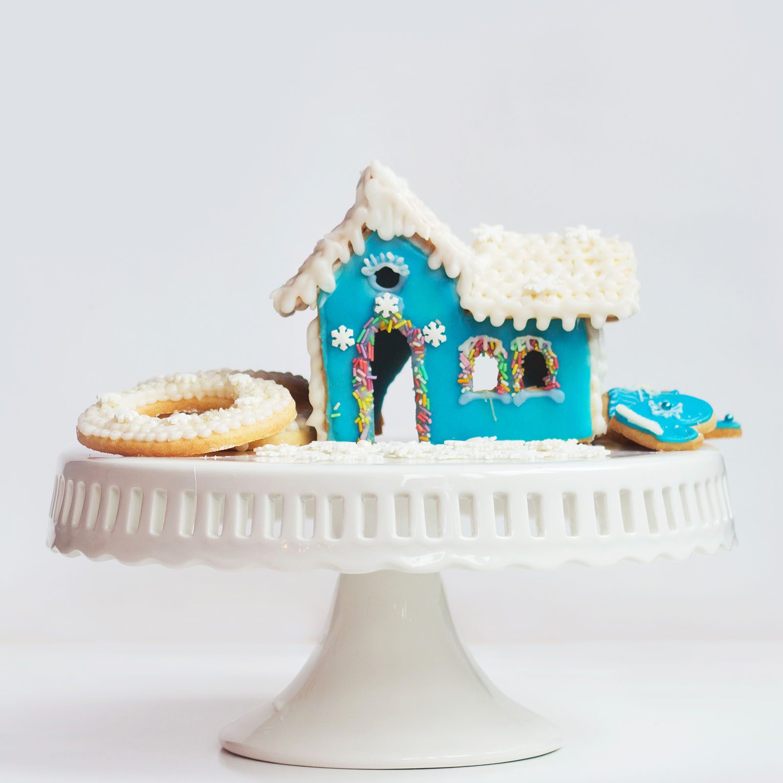 Gingerbread house sits on a white cake stand, decorated with blue icing, sprinkles, and white frosting details, surrounded by frosted cookies.