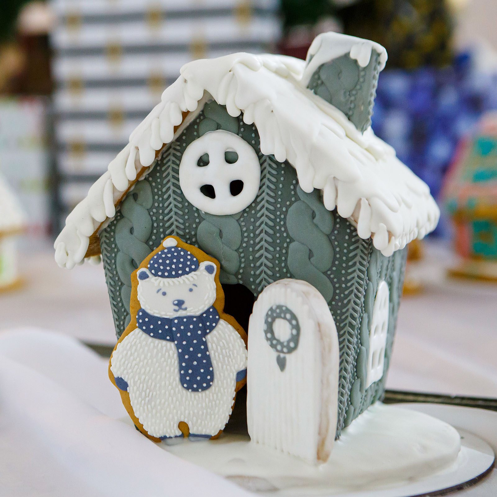 Gingerbread house with frosted roof stands decorated alongside a cookie bear wearing a scarf and hat, set on a festive table.
