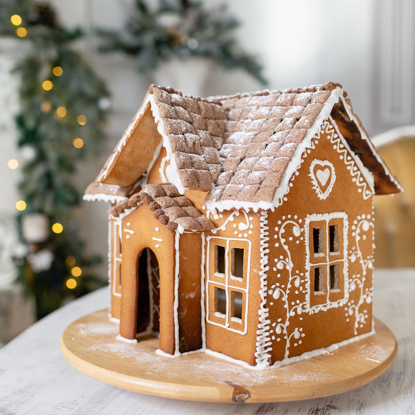 Gingerbread house sits on a wooden plate, decorated with white icing patterns, surrounded by blurred holiday greenery and lights in the background.