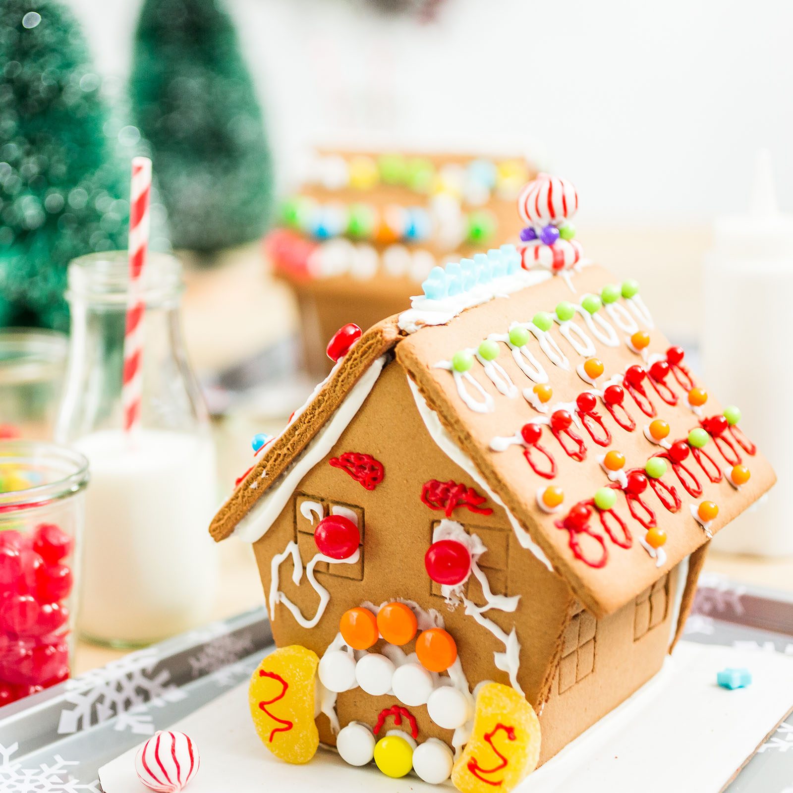 Gingerbread house decorated with colorful candy; sits on a tray with milk and candy jars nearby; miniature Christmas trees in the background.