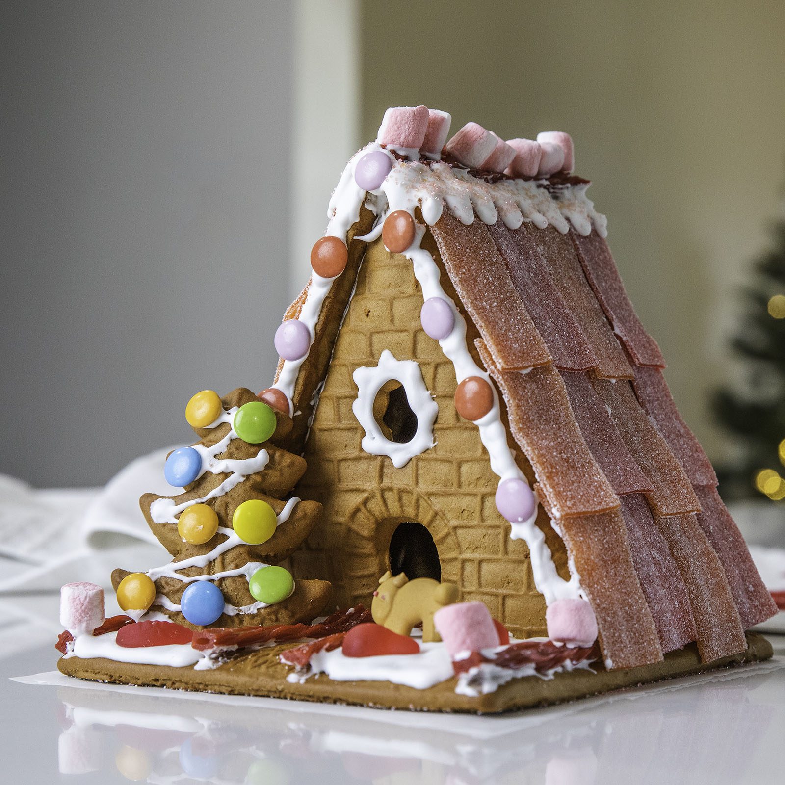 Gingerbread house displaying candies and icing, sits on a table; a decorated cookie tree stands beside it, holiday setting in the background.
