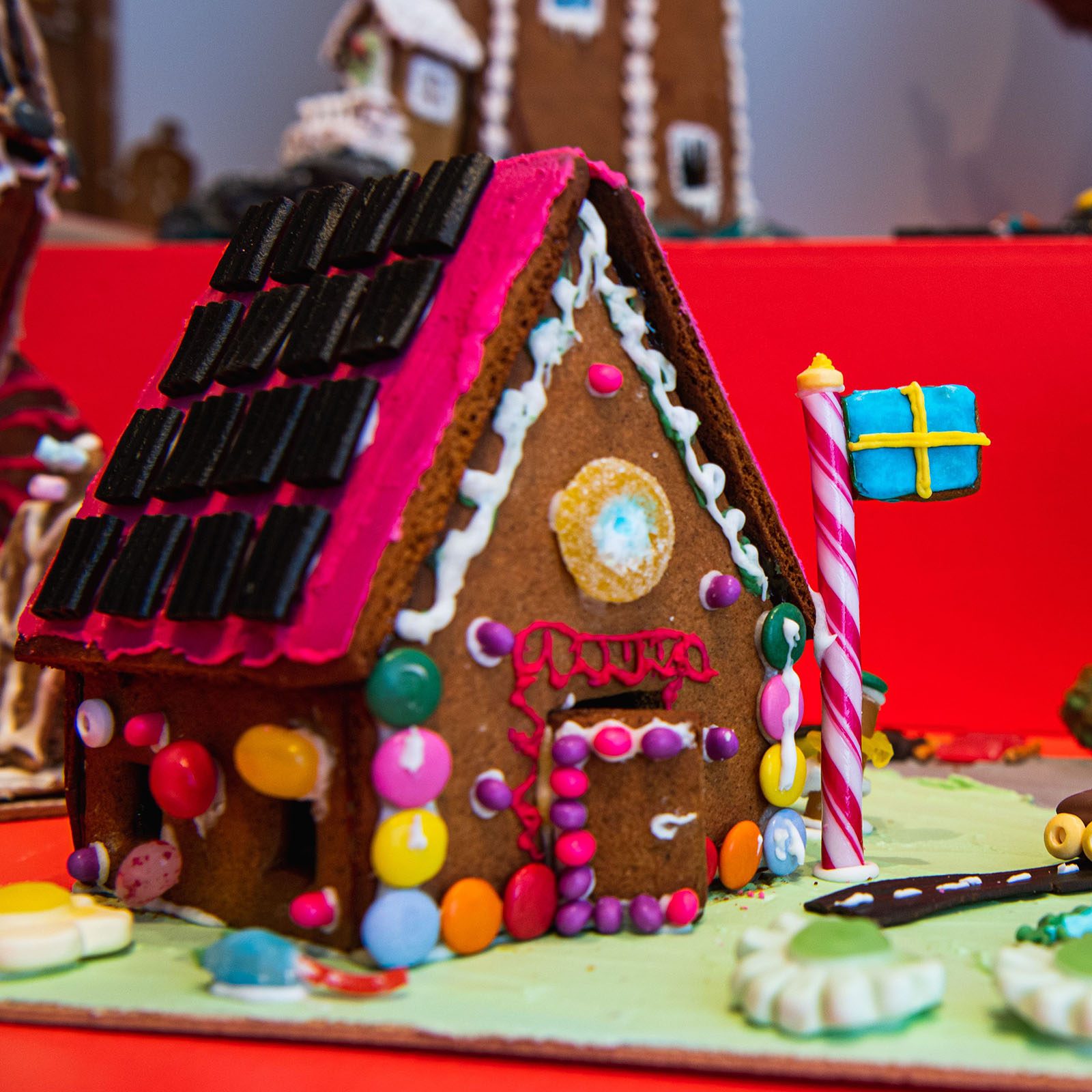 Gingerbread house decorated with candy and licorice, featuring a Swedish flag on a post, set against a bright red background.