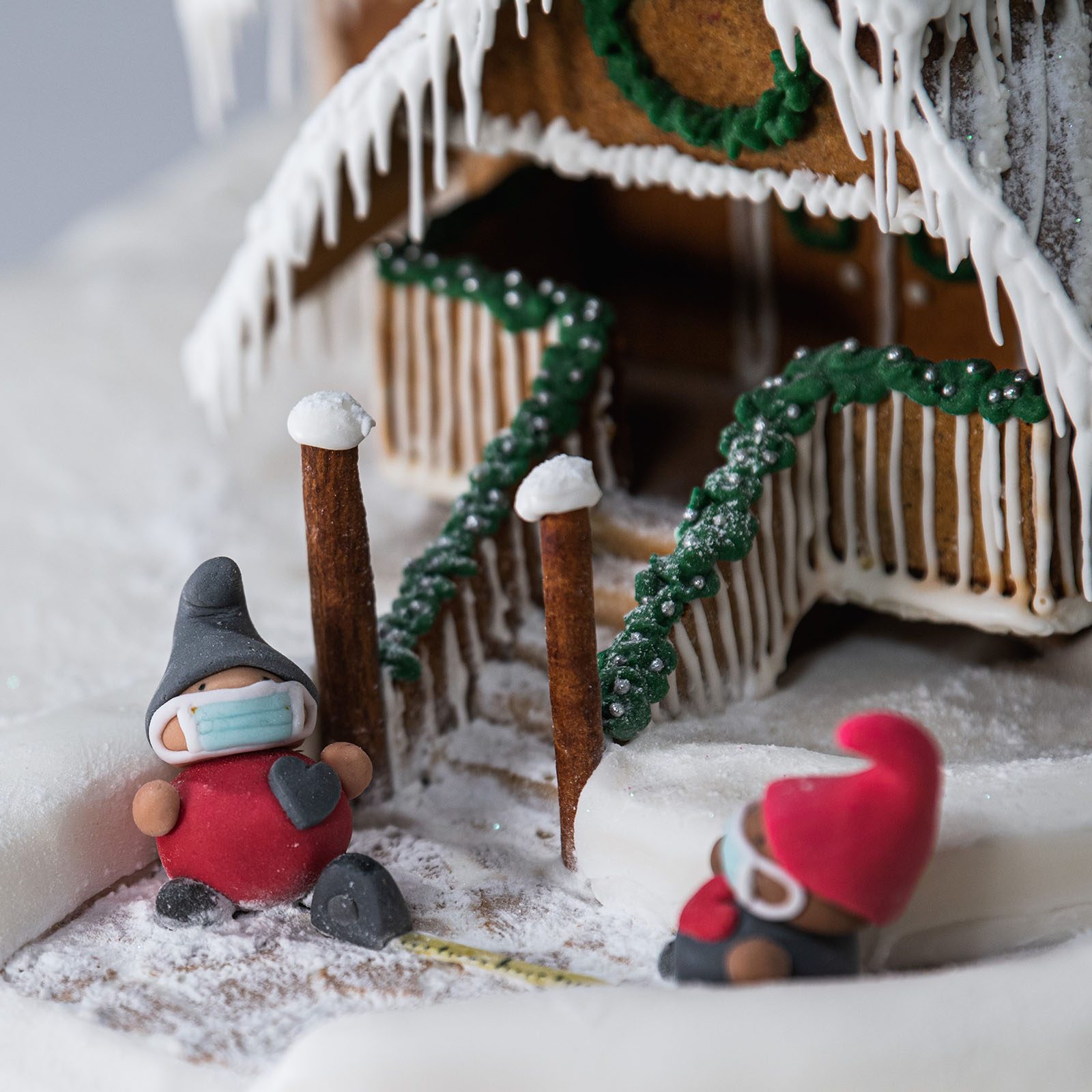 Two masked gnomes stand near a snowy gingerbread house with iced roof, surrounded by decorative green garlands and cinnamon stick trees.