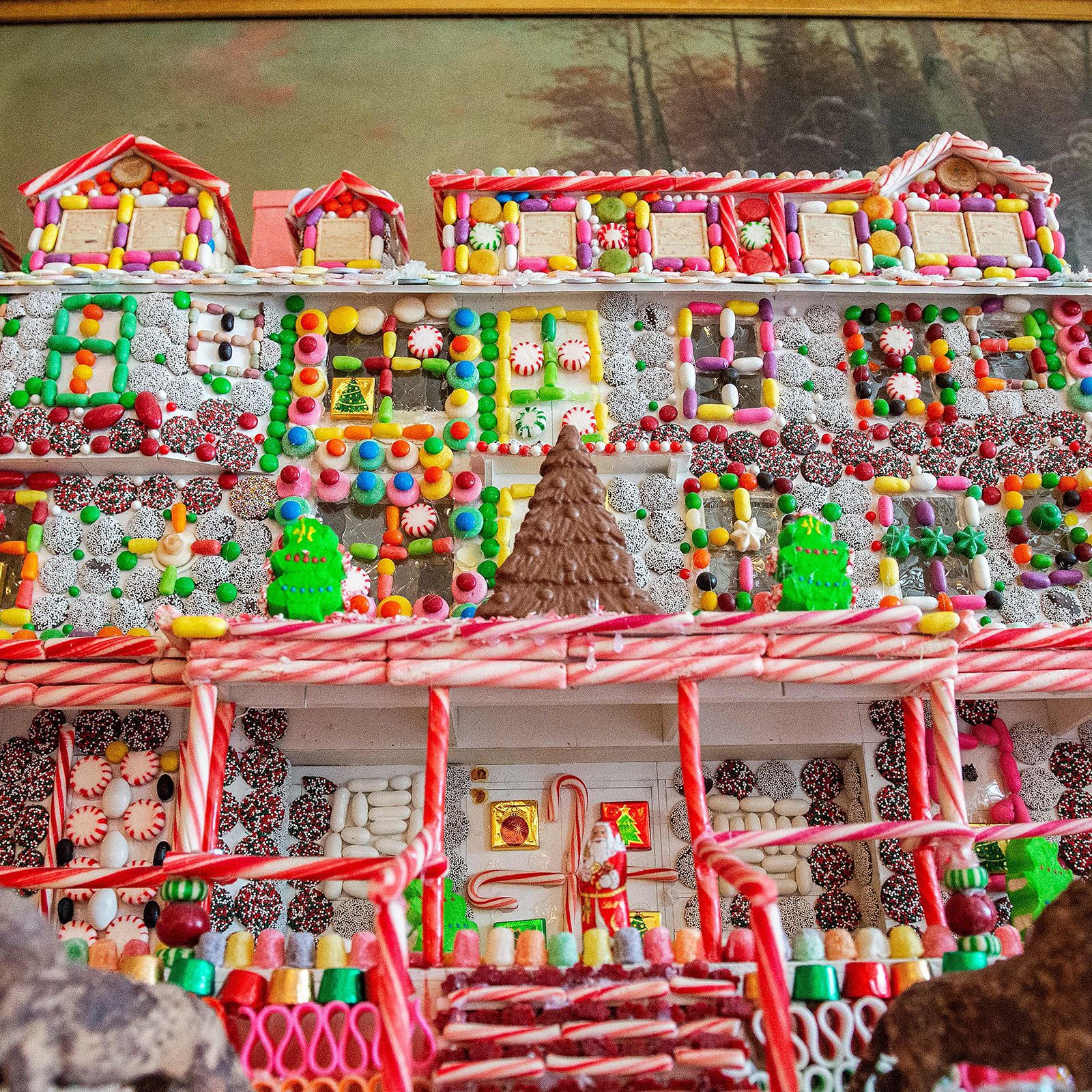 A gingerbread house, adorned with colorful candies, sits on display in a festive setting with a painted landscape in the background.