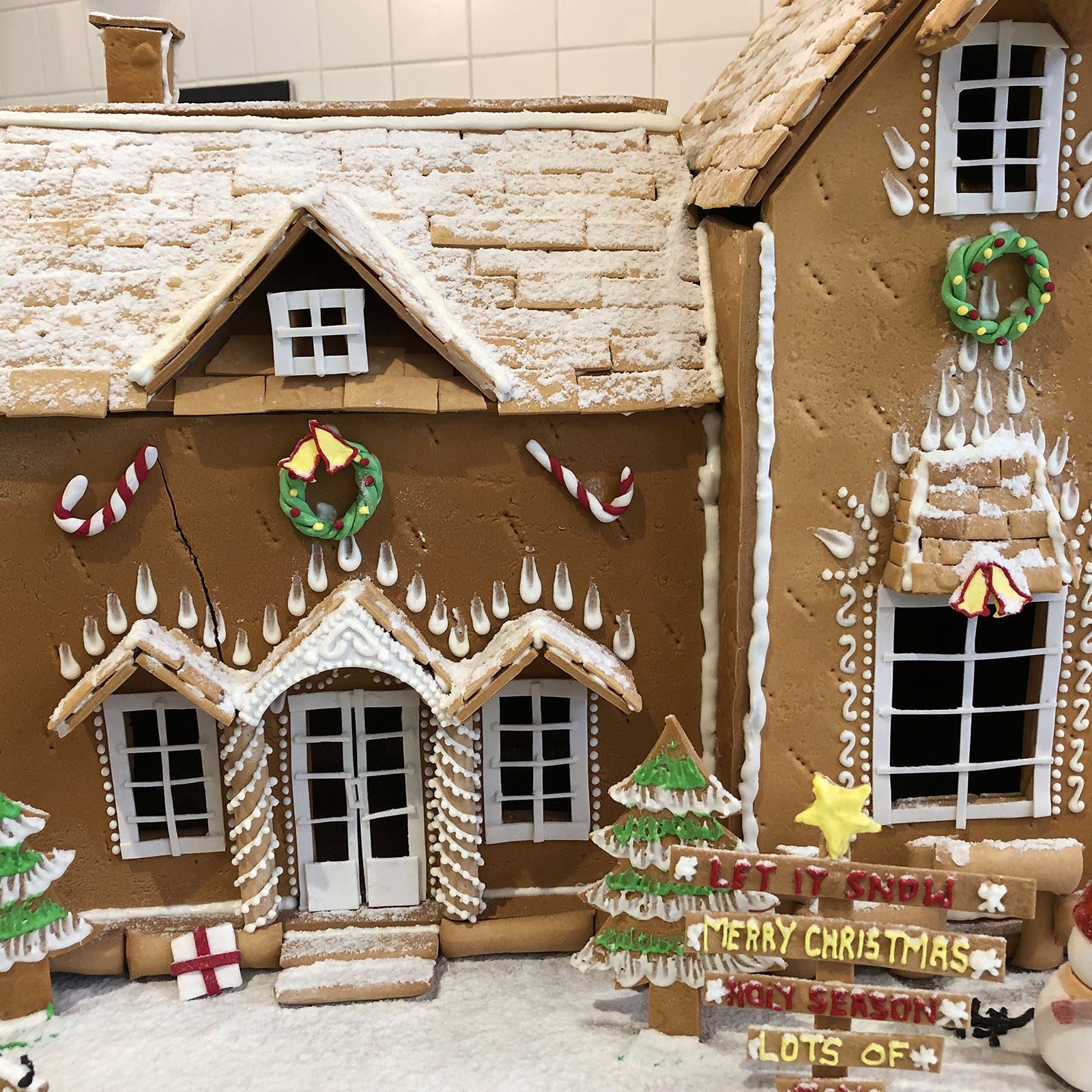 Gingerbread house displays Christmas decorations, including candy canes and wreaths; a sign reads "LET IT SNOW MERRY CHRISTMAS HOLY SEASON LOTS OF."