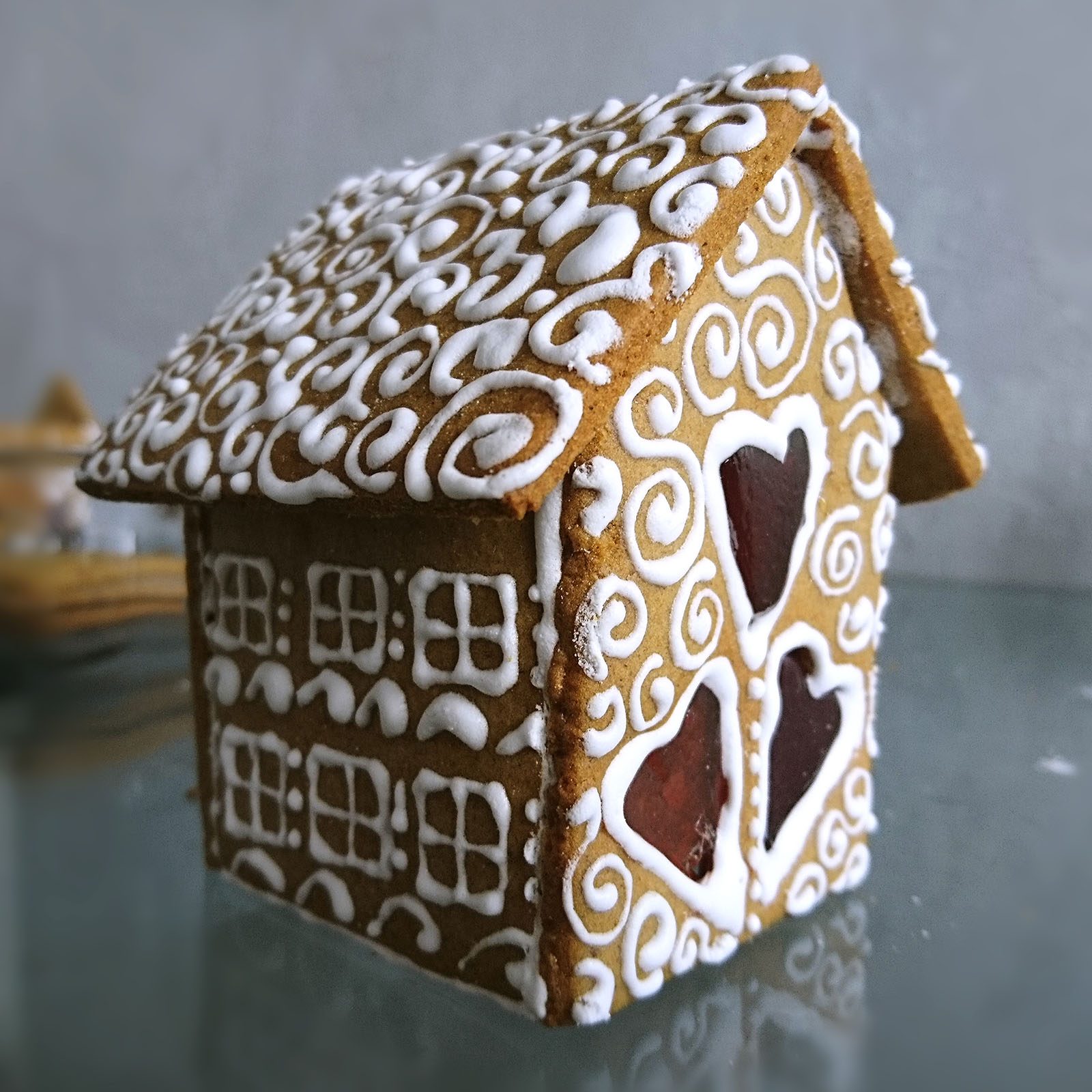 Gingerbread house displays intricate white icing patterns on walls and roof, set on reflective surface, evoking a whimsical, festive atmosphere.