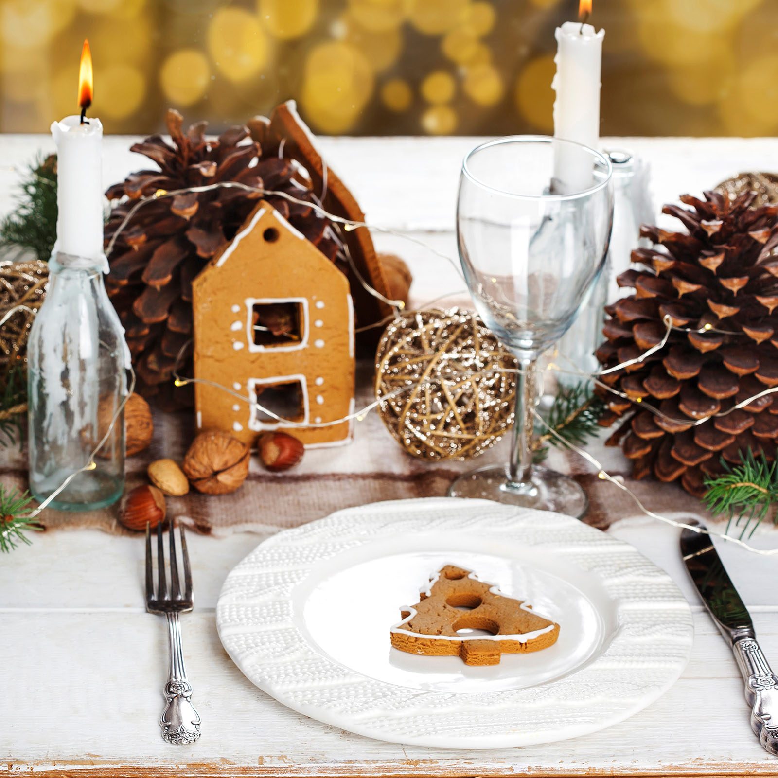 Gingerbread house centerpiece adorns a festive table setting with candles, pinecones, nuts, and a cookie on a plate, surrounded by twinkling lights.