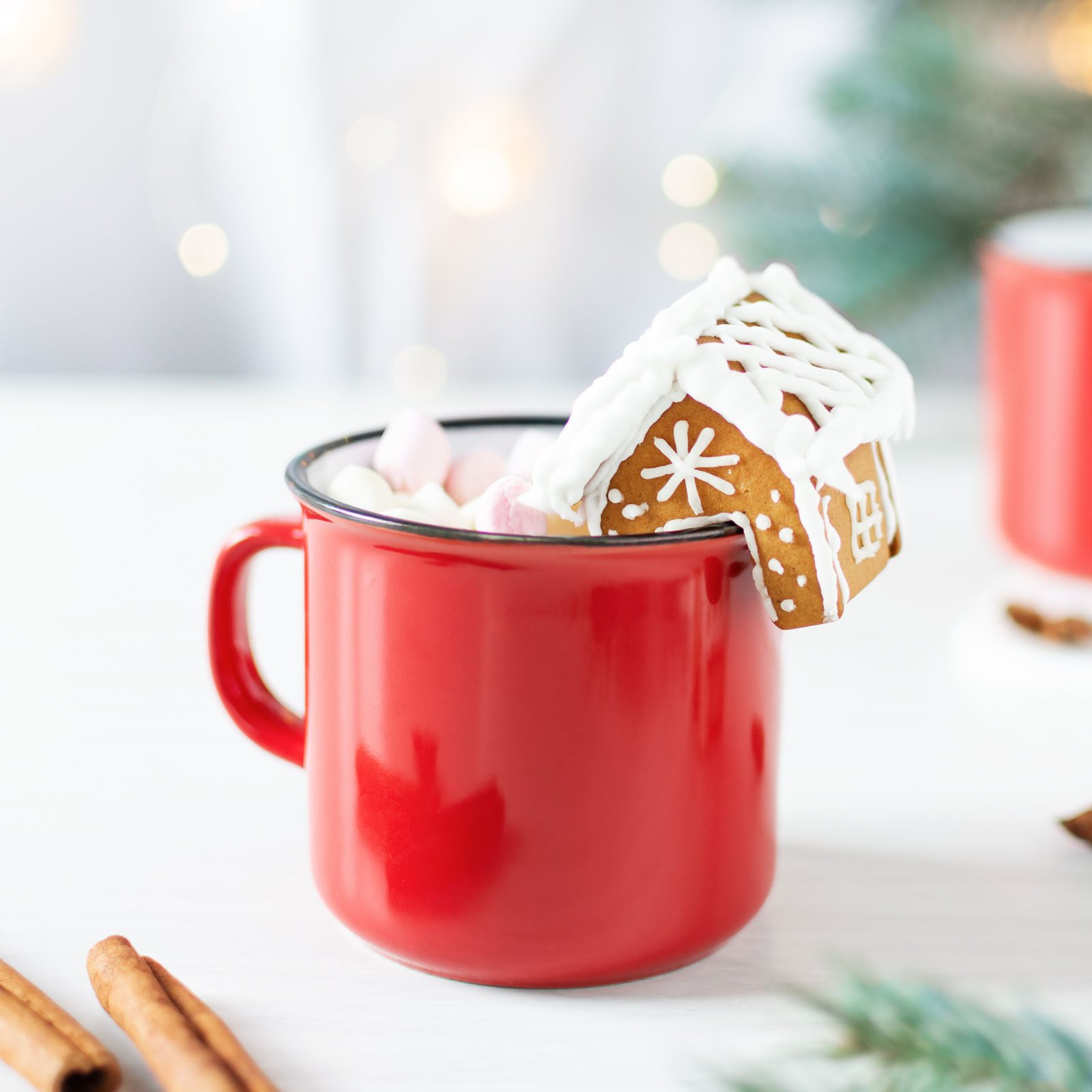 Red mug holding hot chocolate with marshmallows; gingerbread house sits on the rim. Blurred festive background and cinnamon sticks nearby on a white surface.