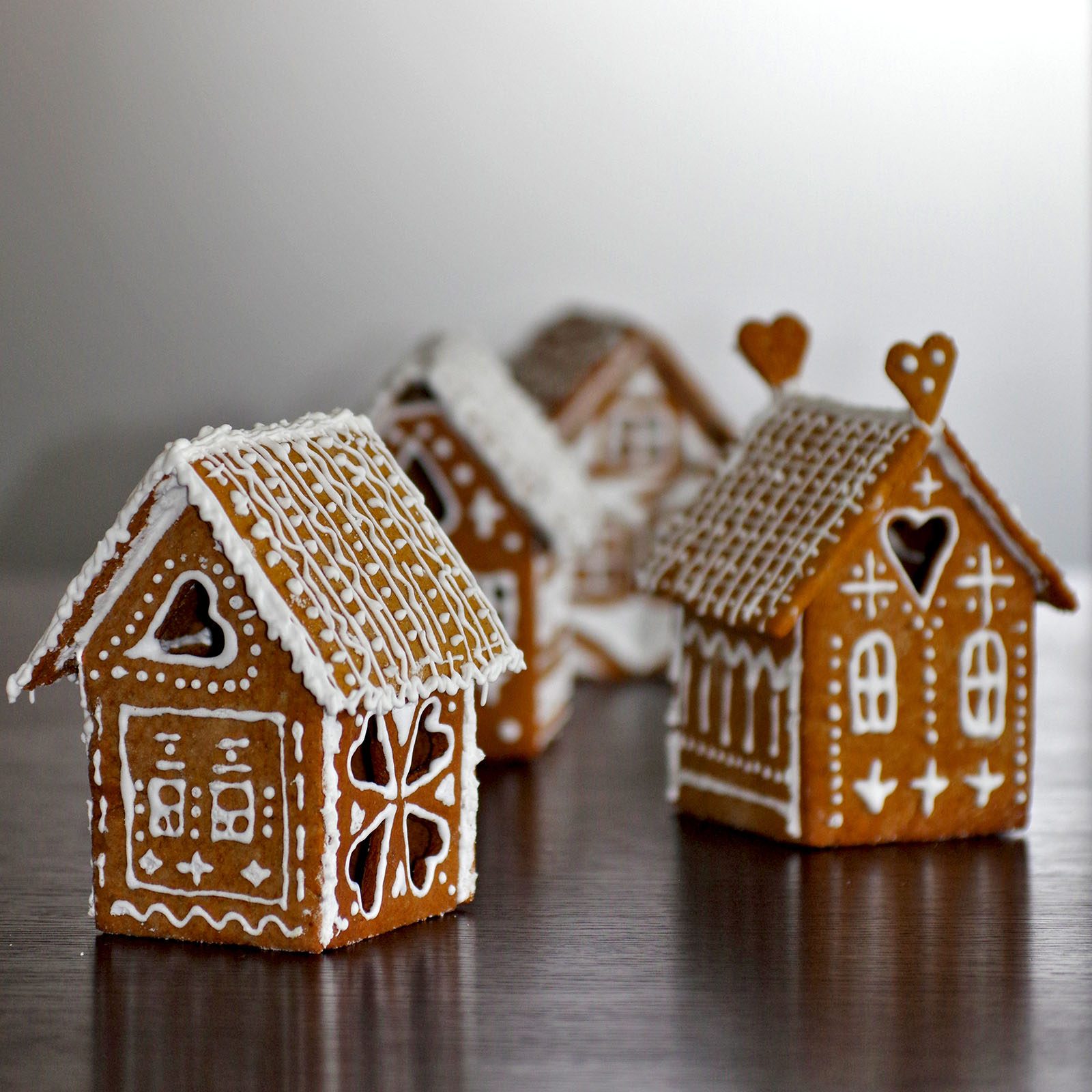 Gingerbread houses decorated with white icing, featuring heart and window designs, sit on a dark surface against a neutral background.