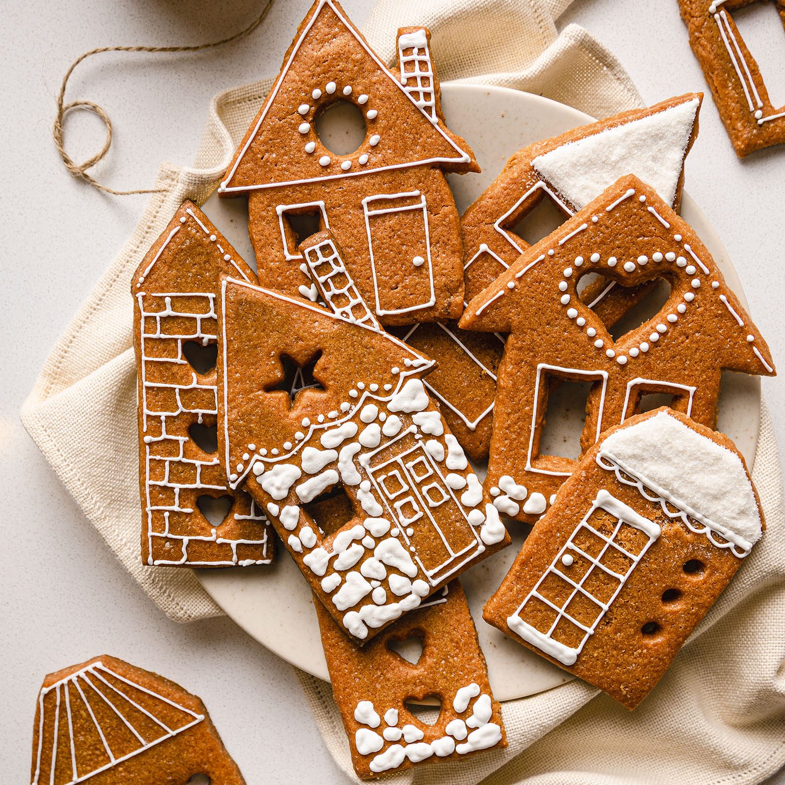 Gingerbread cookies decorated with white icing form house shapes, arranged on a plate, surrounded by a cream cloth on a light surface.