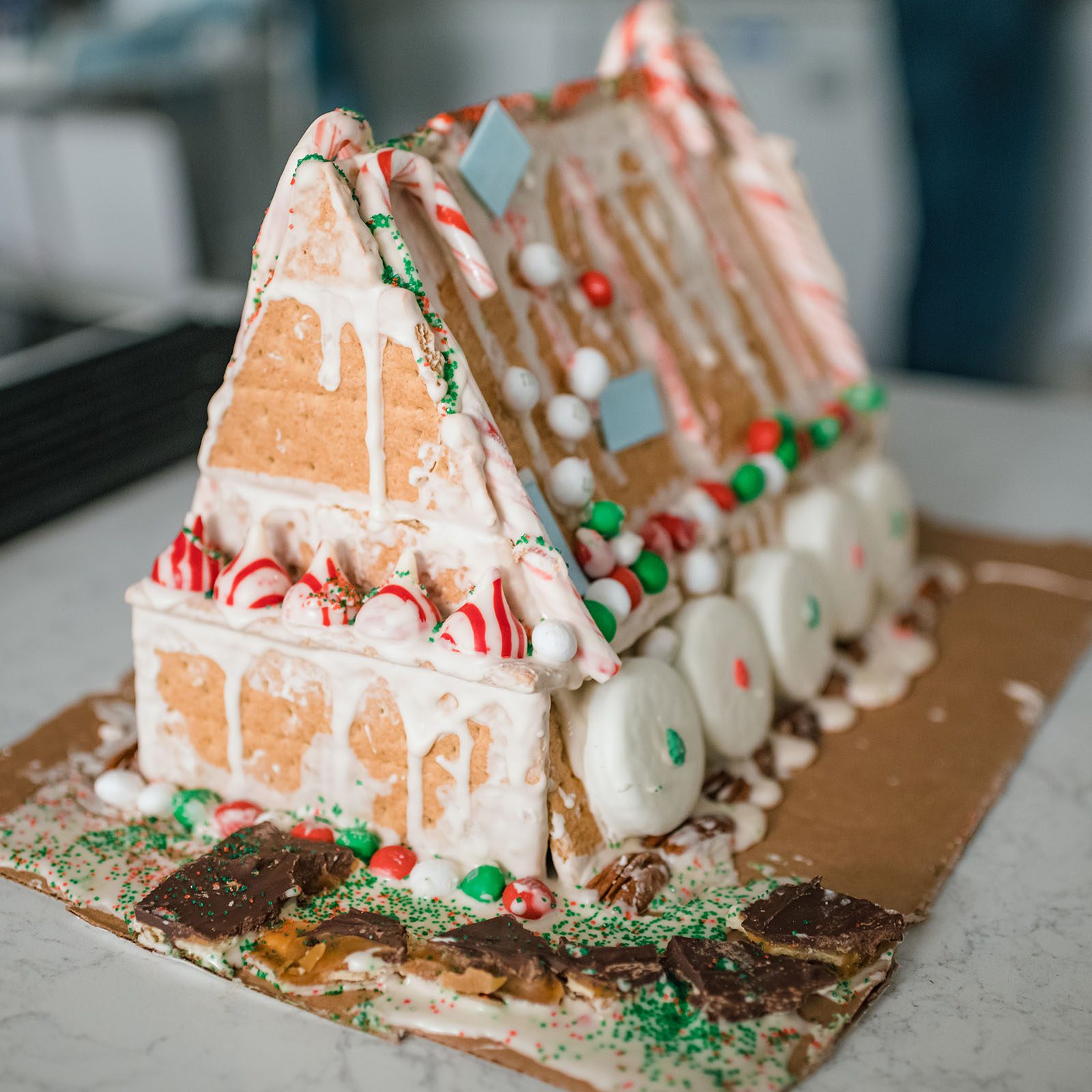 Gingerbread house decorated with icing, candies, and pretzels; sits on a cardboard base with festive sprinkles, on a kitchen counter.