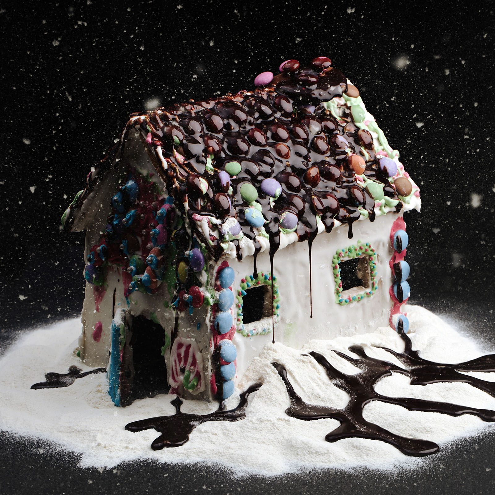 Gingerbread house decorated with colorful candies and chocolate drips, sitting on powdered sugar, with a snowy effect in a dark setting.