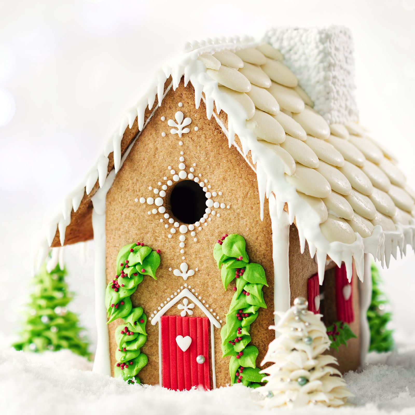 Gingerbread house adorned with icing and candy, sitting in snow, surrounded by small Christmas trees, conveying a festive holiday atmosphere.