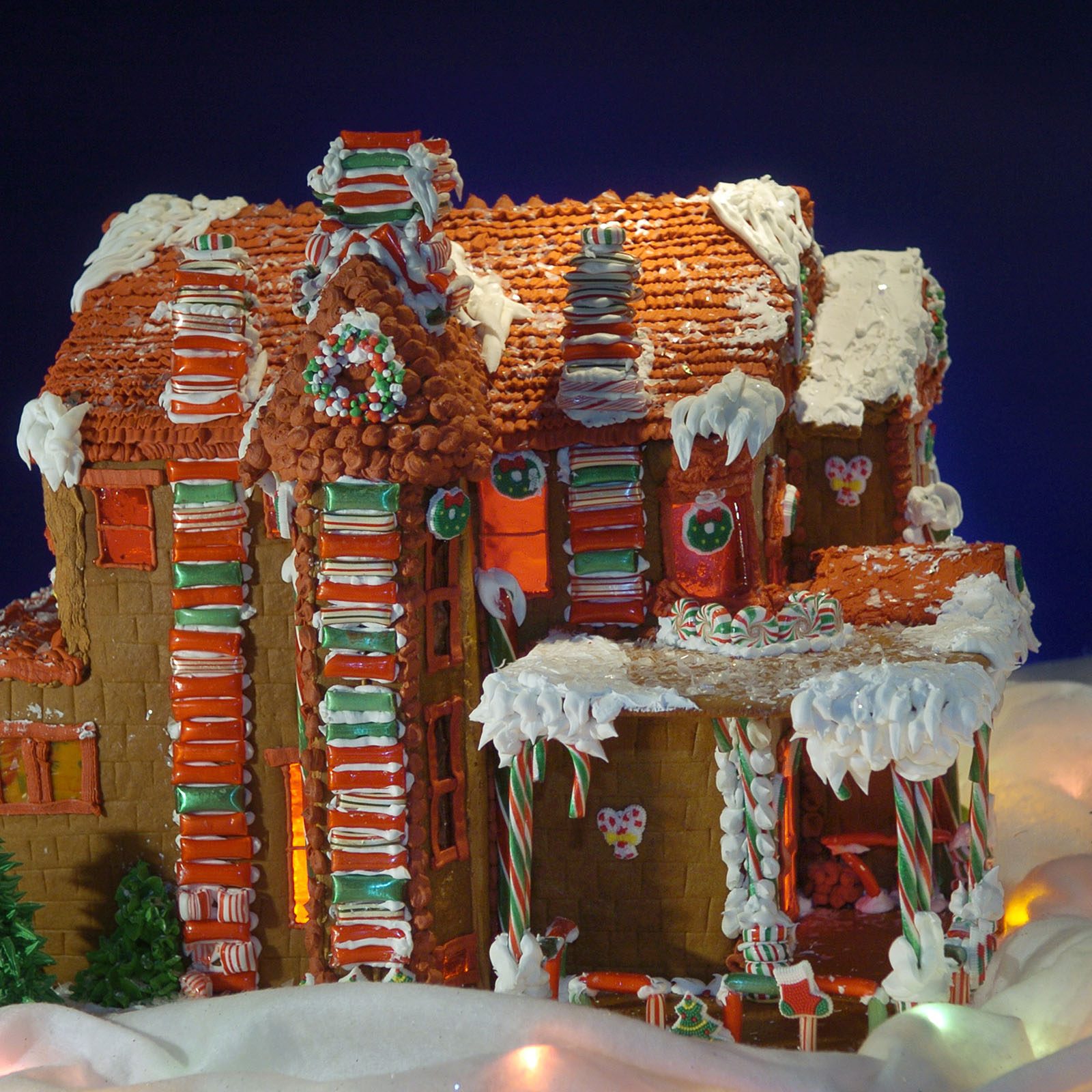 Gingerbread house displays colorful candy decorations amidst icing snow, creating a festive holiday scene against a dark blue background.