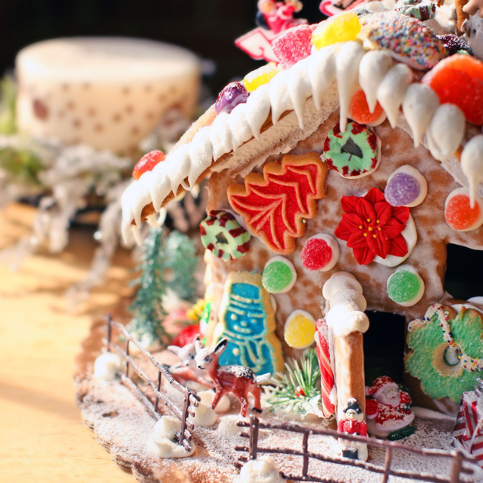 Gingerbread house adorned with colorful candies sits on a snowy base, surrounded by festive miniatures and rustic fence, evoking a holiday theme.