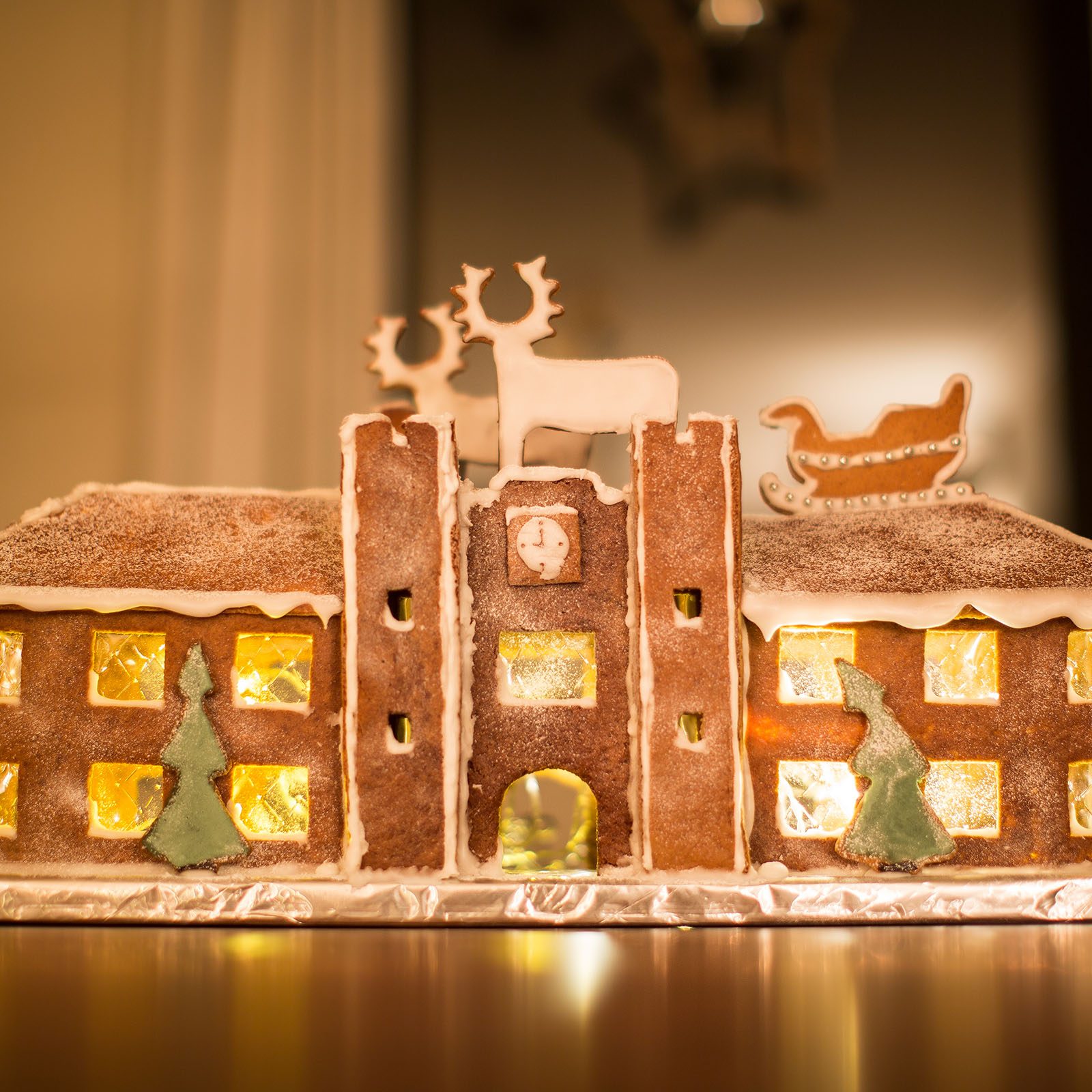 Gingerbread house displays glowing windows, topped with icing reindeer and sleigh, set on a reflective surface in a warmly lit room.