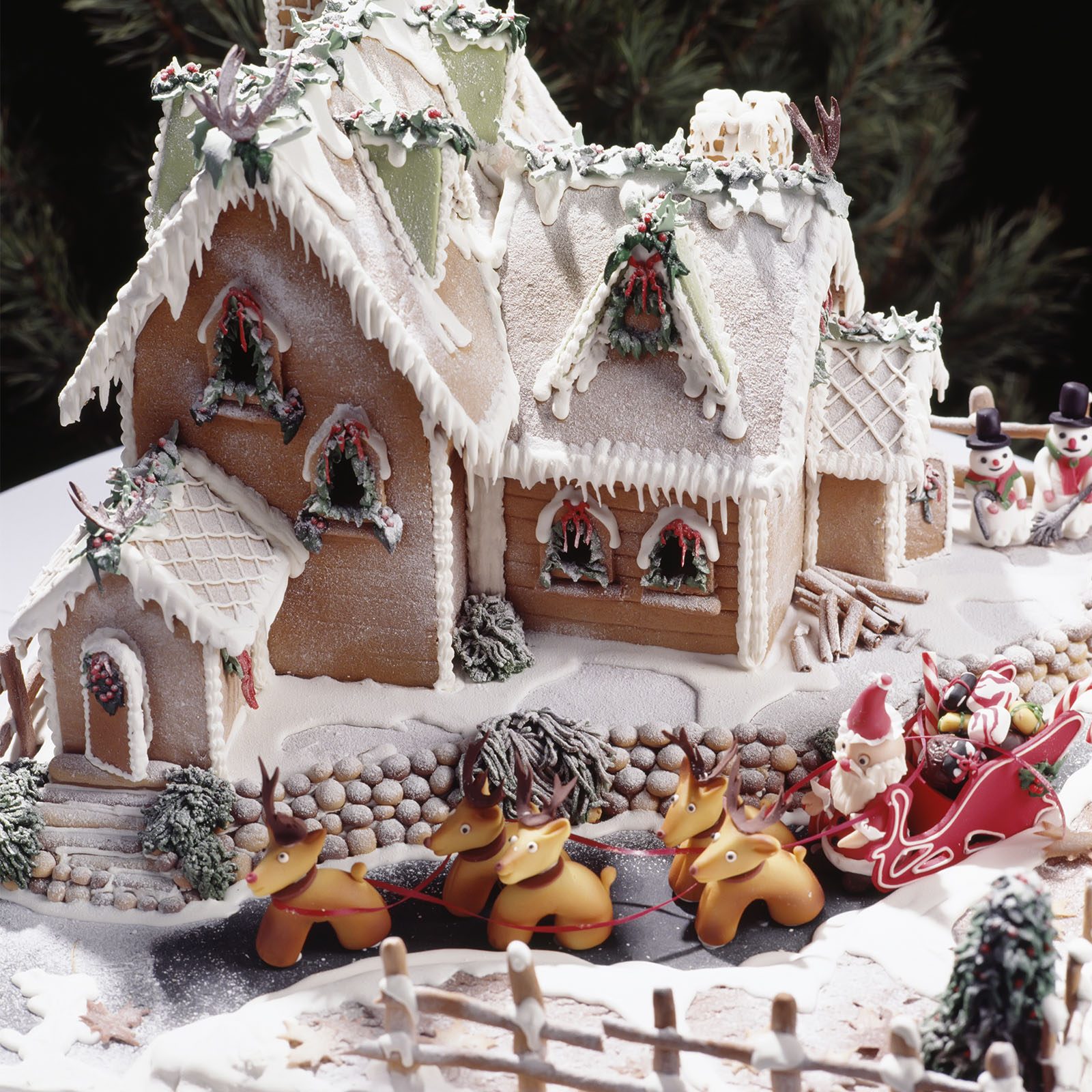 Gingerbread house sits amidst icing snow; Santa's sleigh led by reindeer nearby; snowmen in background, adorned with festive decorations.