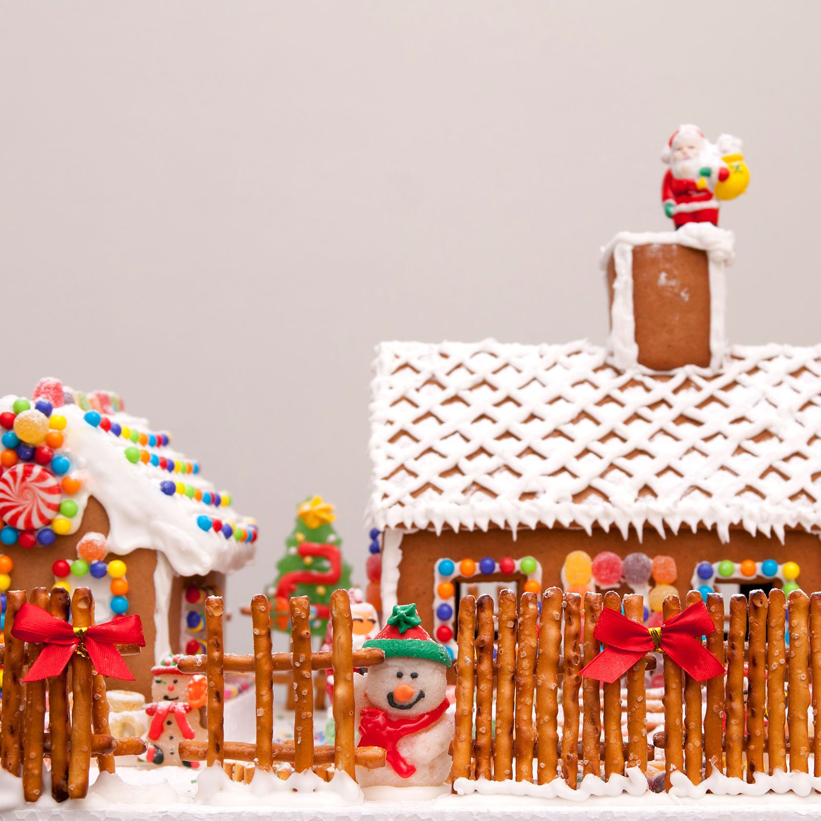 Gingerbread house adorned with candy sits behind a pretzel fence. Snowman figure stands by, Santa decoration atop the chimney, festive holiday scene.