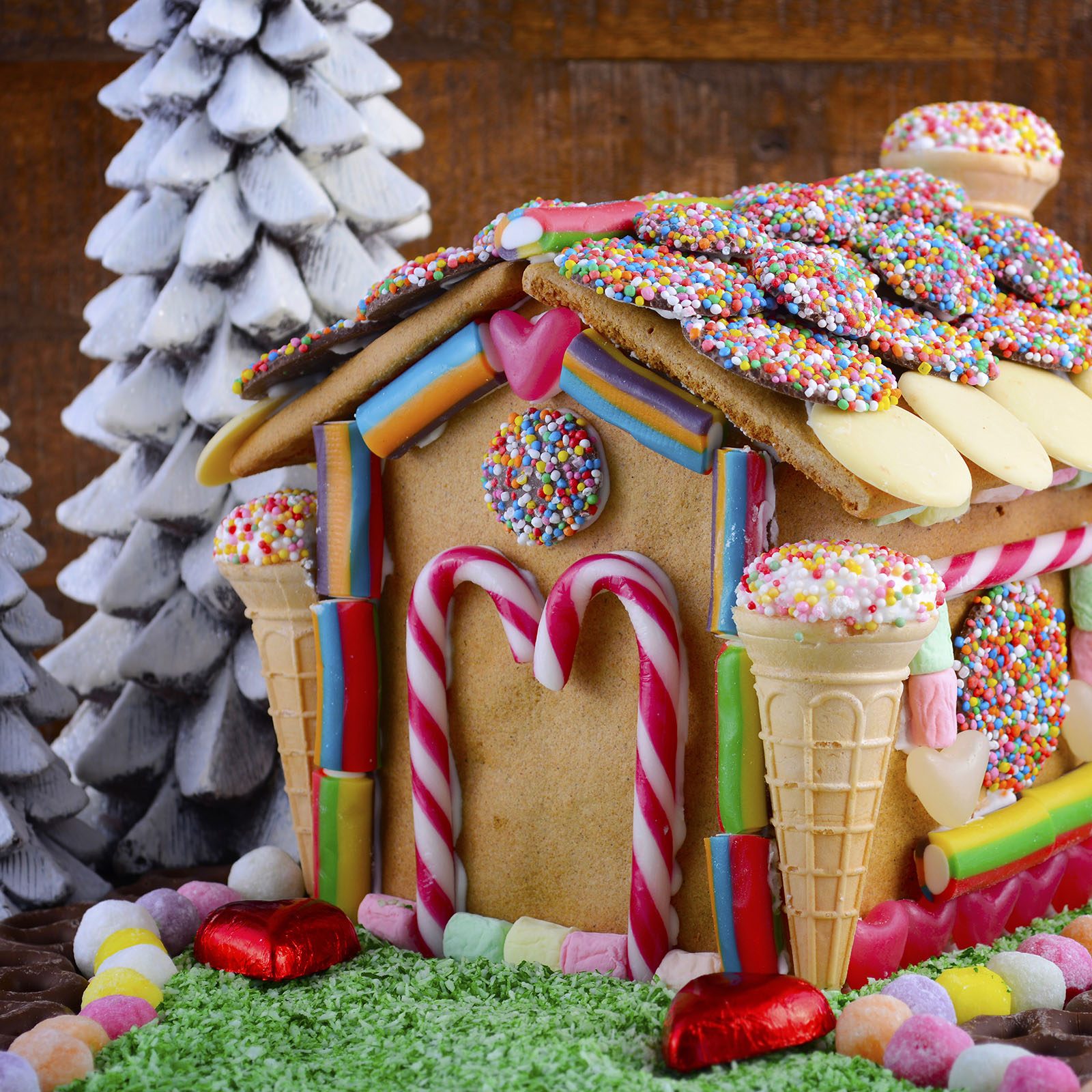 Gingerbread house displays colorful candy decorations; surrounded by snowy trees, it sits on a green sugary lawn.