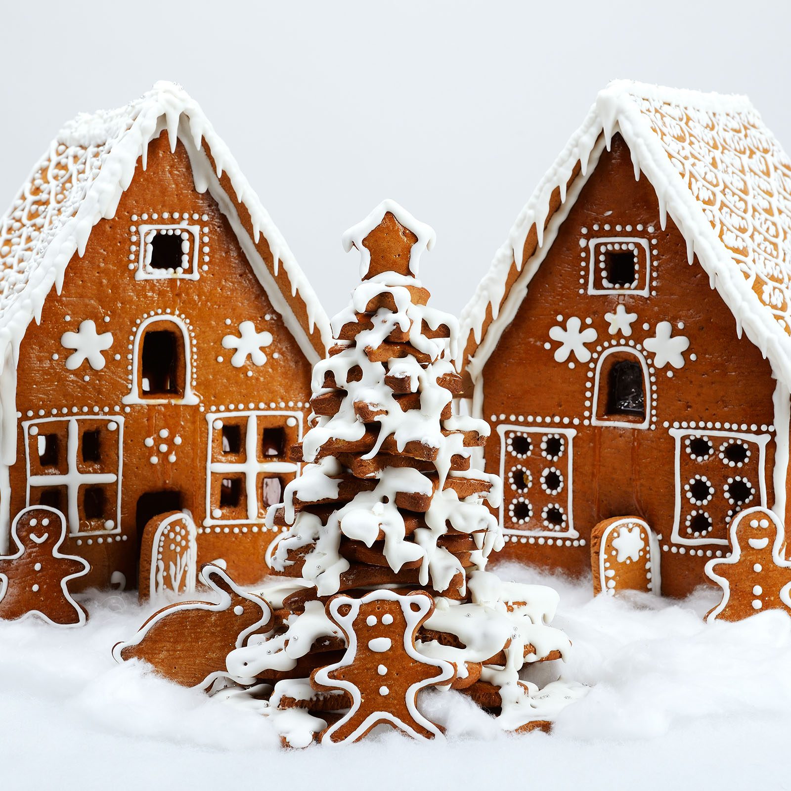 Gingerbread houses stand decorated with white icing, accompanied by gingerbread cookies and icing-covered tree, set on cotton-like snow.