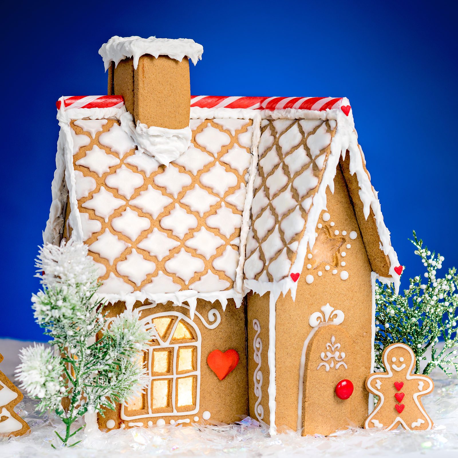 Gingerbread house stands festively adorned with icing and candy, surrounded by small frosted trees on a snowy surface against a blue background.