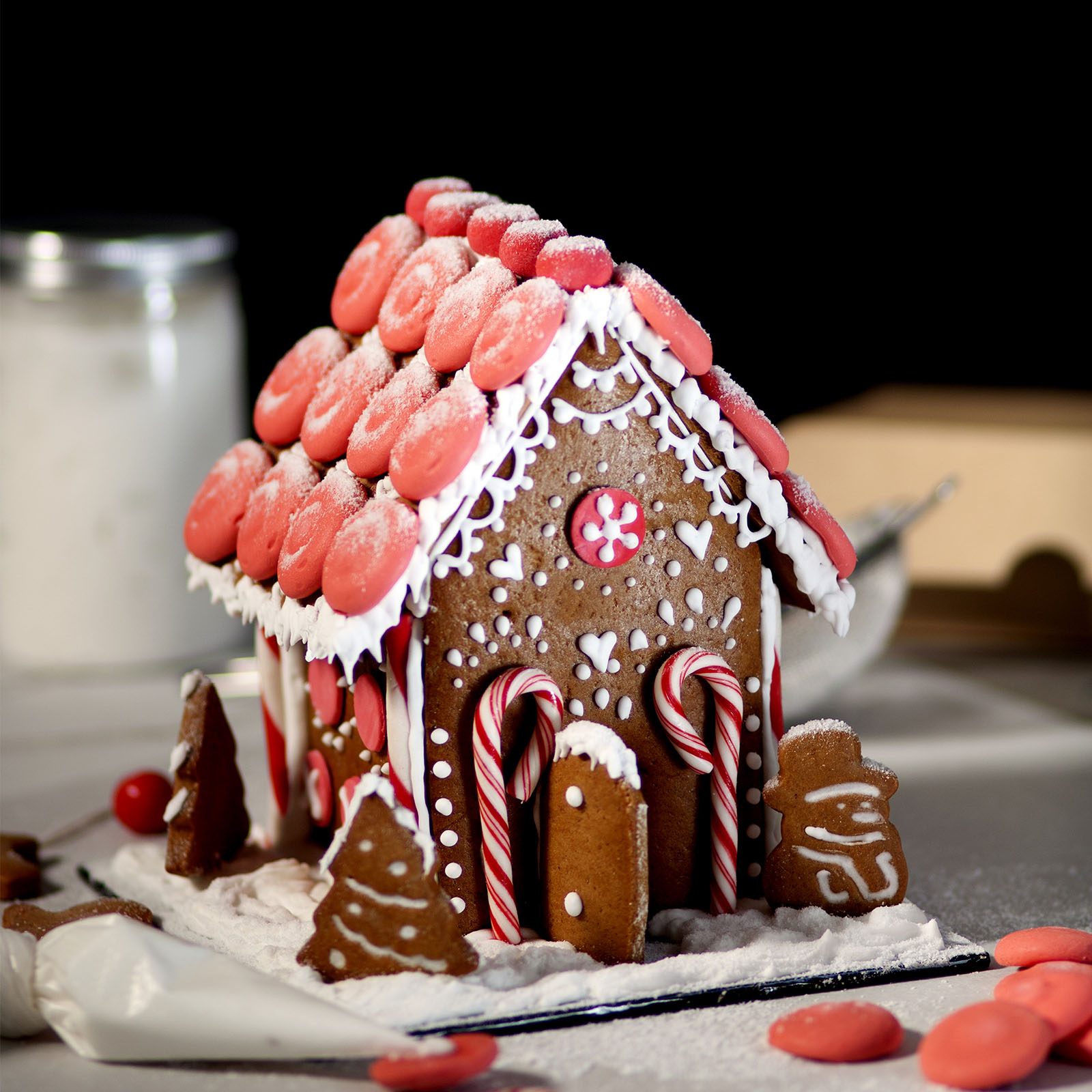 Gingerbread house adorned with red candies and icing, surrounded by cookie trees and candies, sitting on a snowy surface with a dark background.