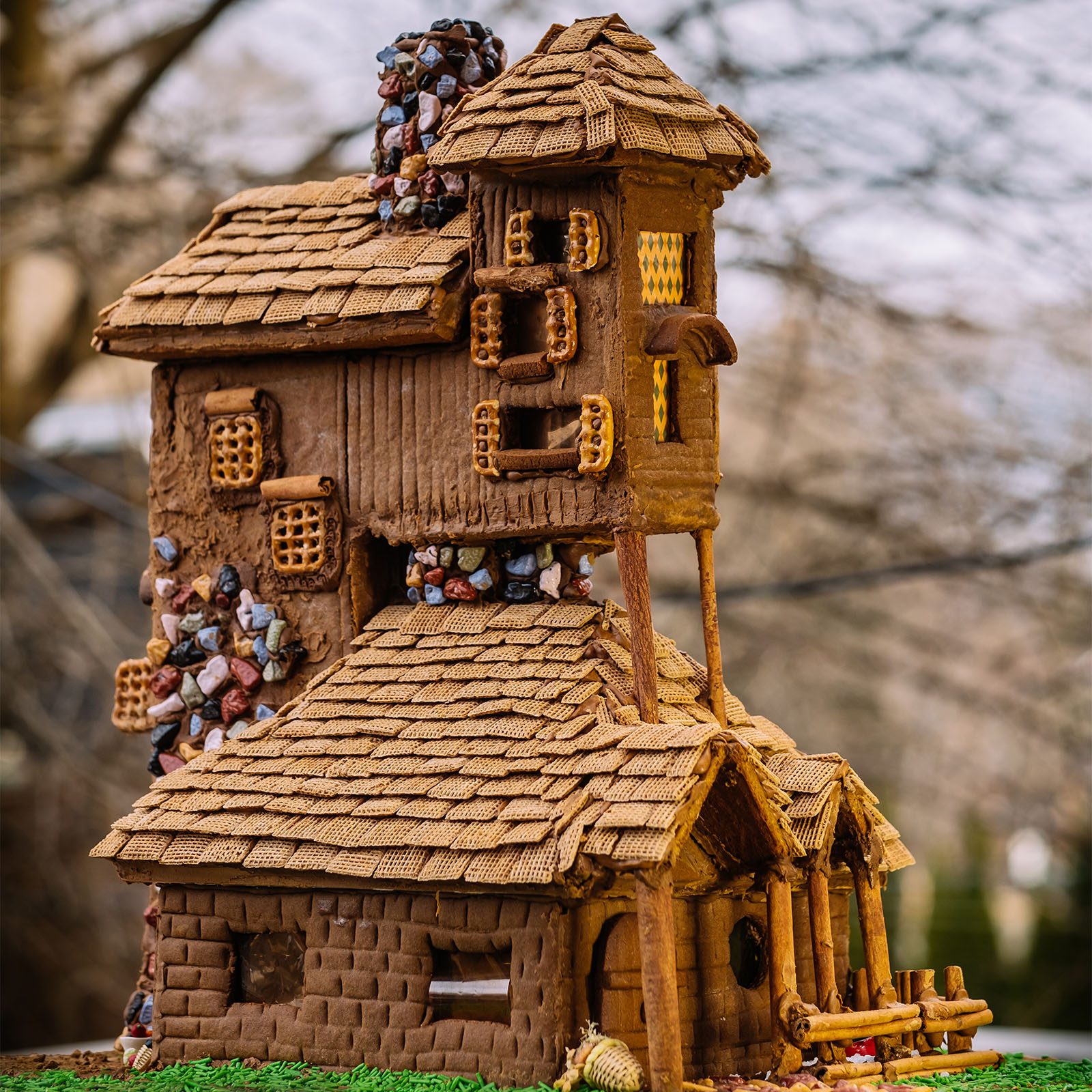 Intricate gingerbread house stands on grass, featuring pretzel windows and candy rocks, with a multi-roof design amidst a blurred tree background.