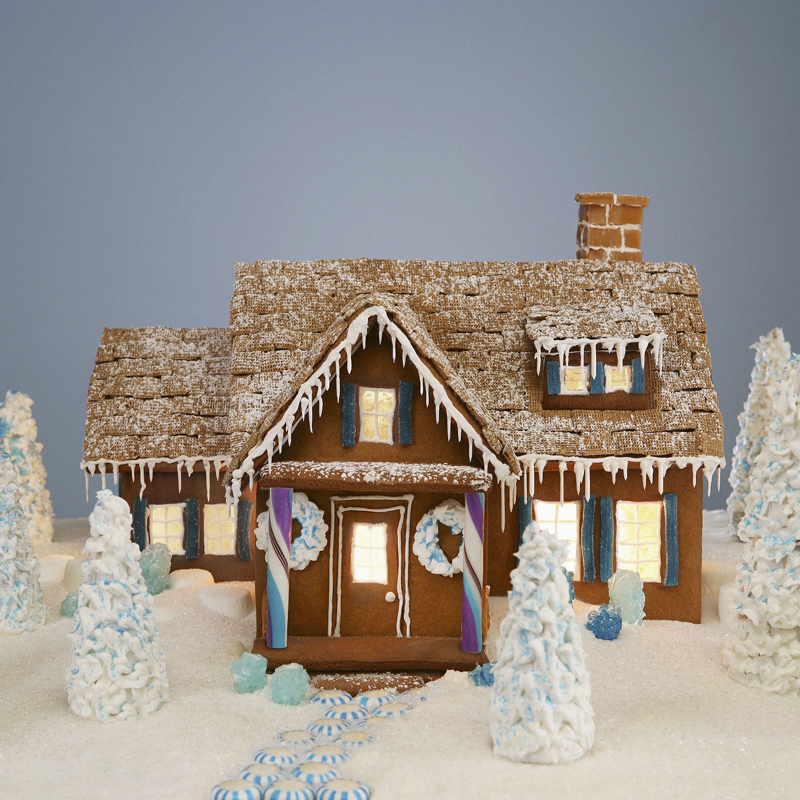 Gingerbread house stands in a snowy setting, adorned with icing icicles, peppermint pathway, and candy-detailed trees under a clear, blue sky.