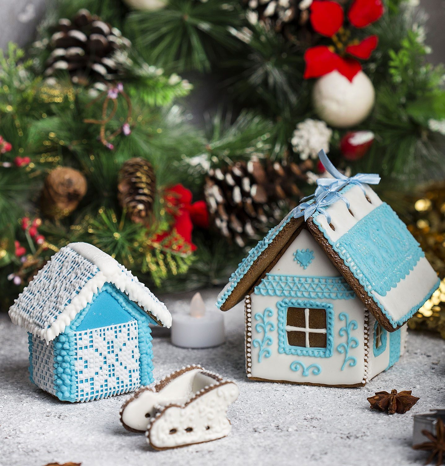 Gingerbread houses decorated with blue and white icing resting on a snowy surface, surrounded by evergreen branches and pinecones.