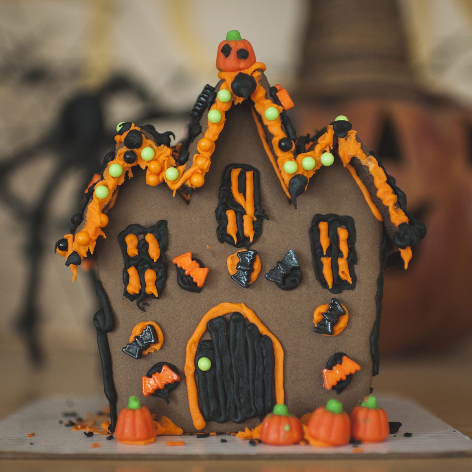A gingerbread haunted house decorated with orange and black icing stands on a table, surrounded by small candy pumpkins in a Halloween-themed setting.