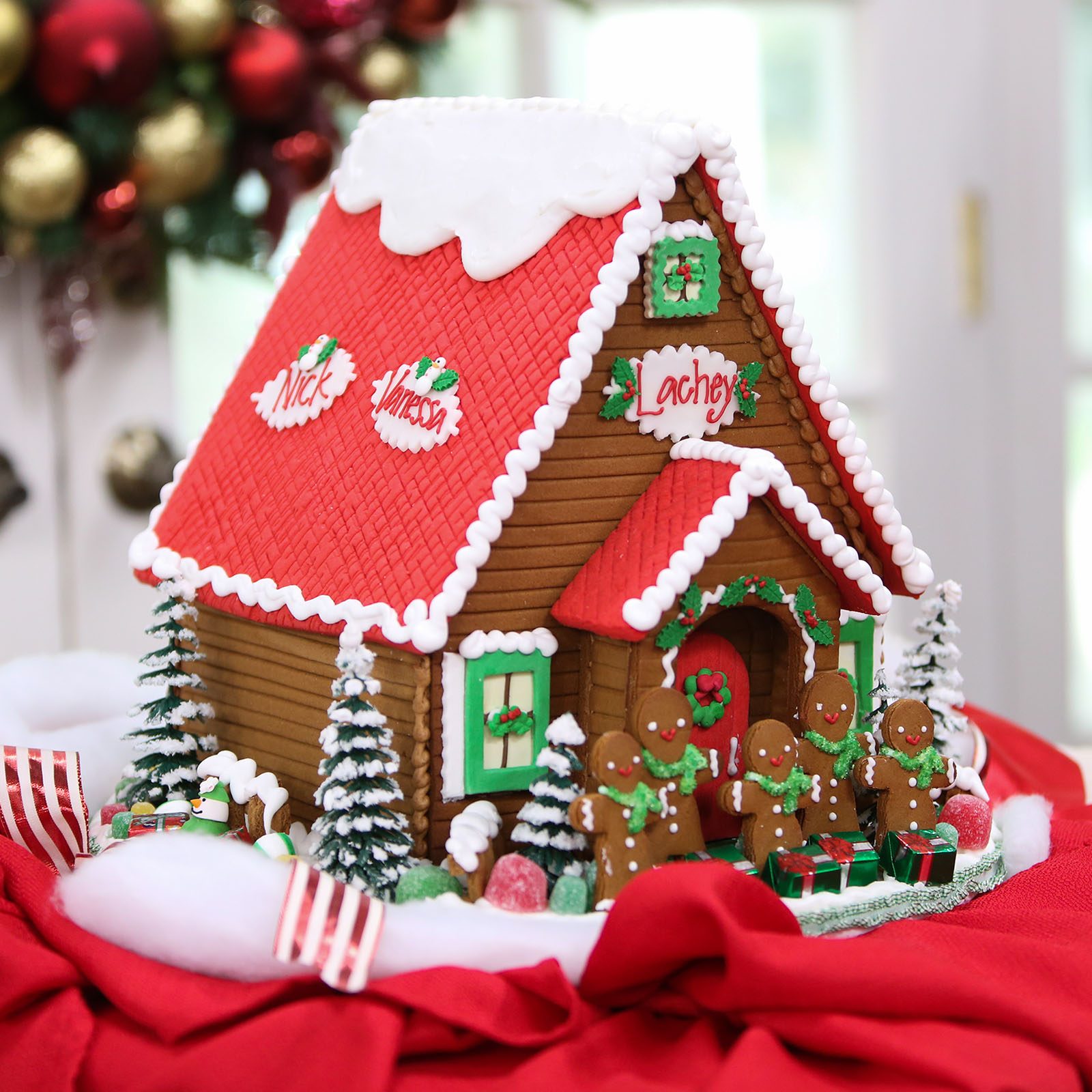 Gingerbread house decorated with icing and candy, surrounded by gingerbread figures, set on a red cloth, with festive ornaments and backdrop. Text: "Nick", "Vanessa", "Lachey".
