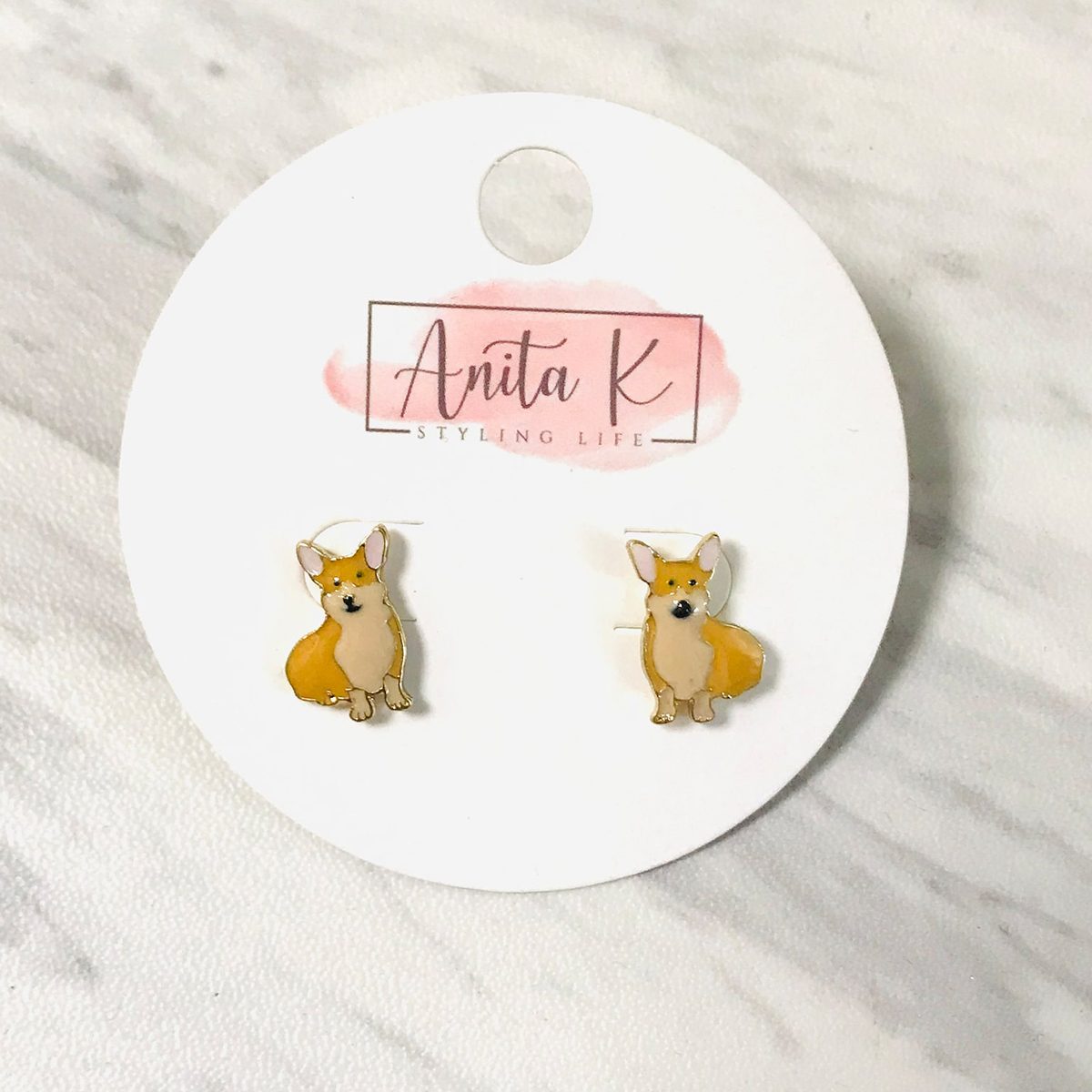 Corgi earrings rest on a round card labeled "Anita K Styling Life," placed on a marbled surface.