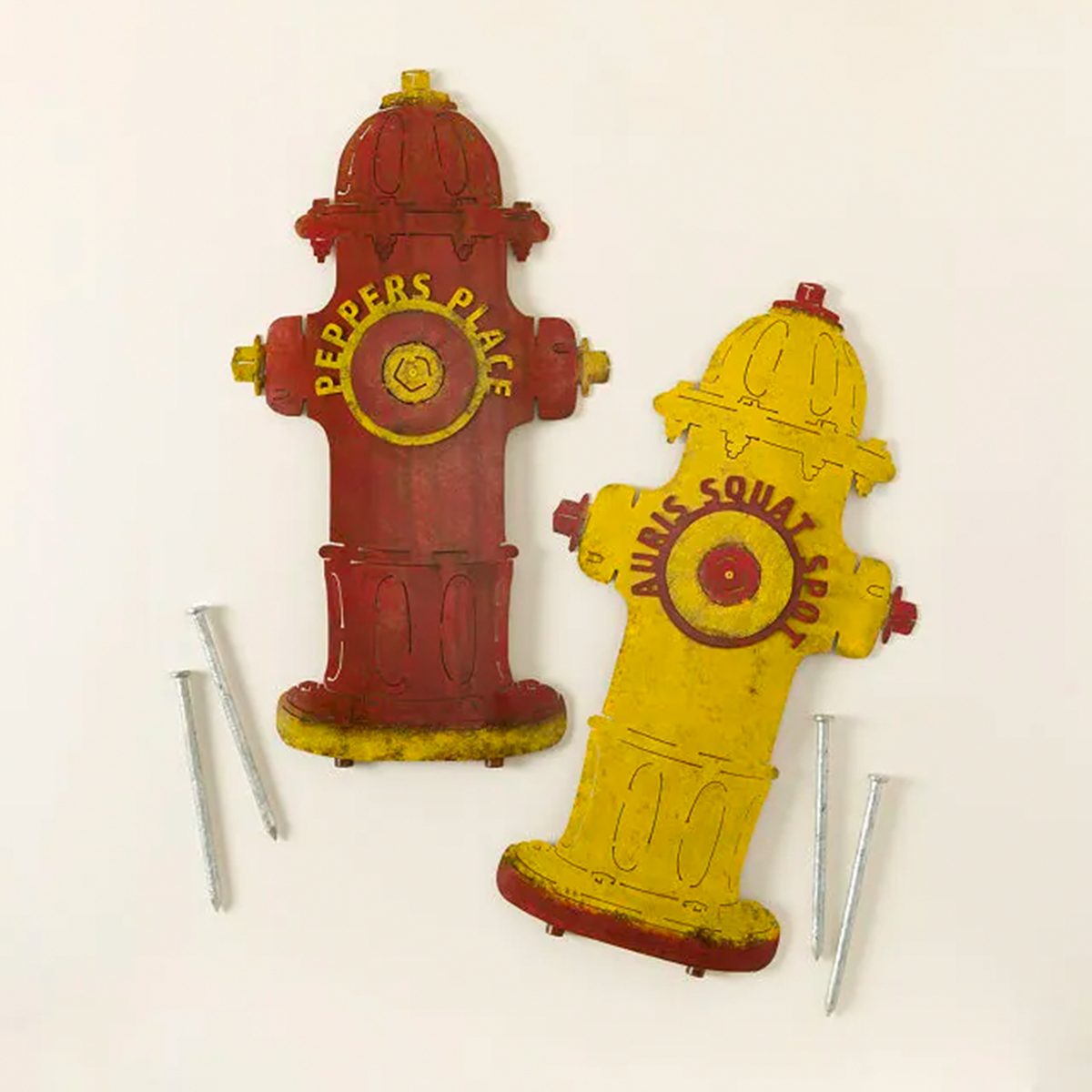 Two cutout fire hydrants, one red and one yellow, labeled "PEPPER'S PLACE" and "ALWAYS SQUAT SPOT," lie next to four nails on a white surface.