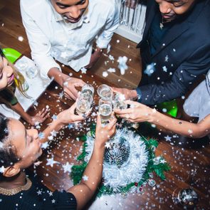 People clink champagne glasses, celebrating around a festive table with tinsel and a disco ball, surrounded by falling star confetti.