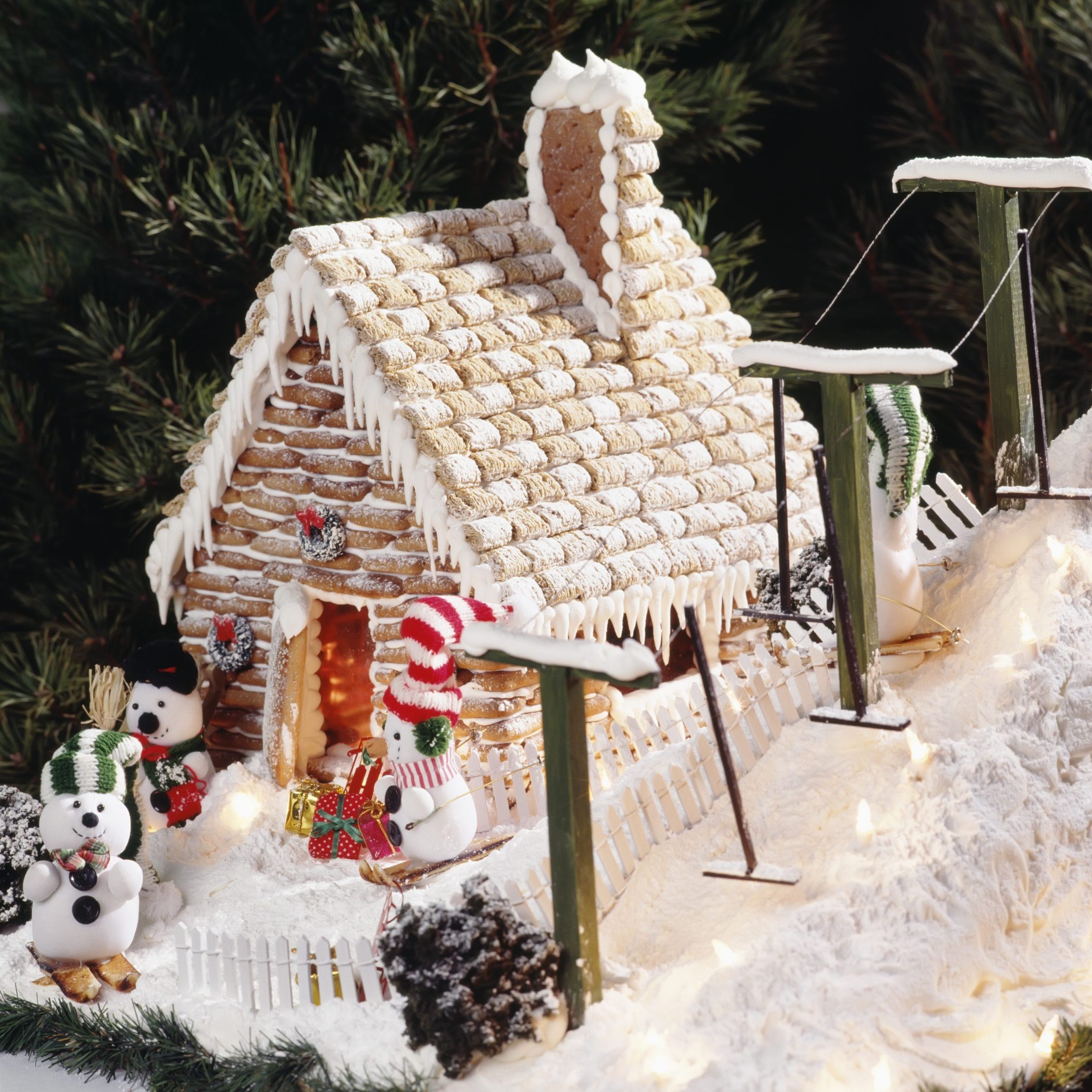 Gingerbread house sits adorned with icing, surrounded by snowman figurines and a snowy landscape, with trees in the background and soft lights illuminating.