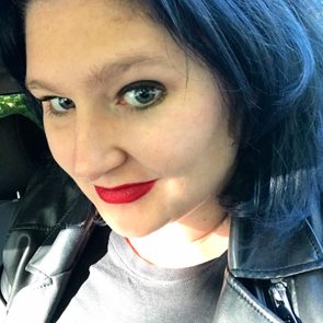 A person with dark hair and red lipstick smiles slightly, wearing a black leather jacket, seated inside a vehicle with sunlight filtering through.