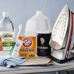 Iron resting on ironing board with vinegar, toothbrush, baking soda, toothpaste, salt, and cleaning cloth, set against a textured blue wall.