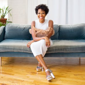 Person smiling, sitting on a blue sofa, wearing a white dress and silver shoes in a room with plants and wooden floor.