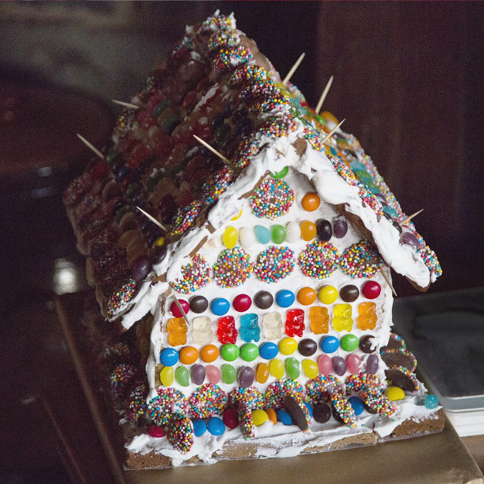 Gingerbread house decorated with colorful candies and sprinkles, resting on a table in a dimly lit room.