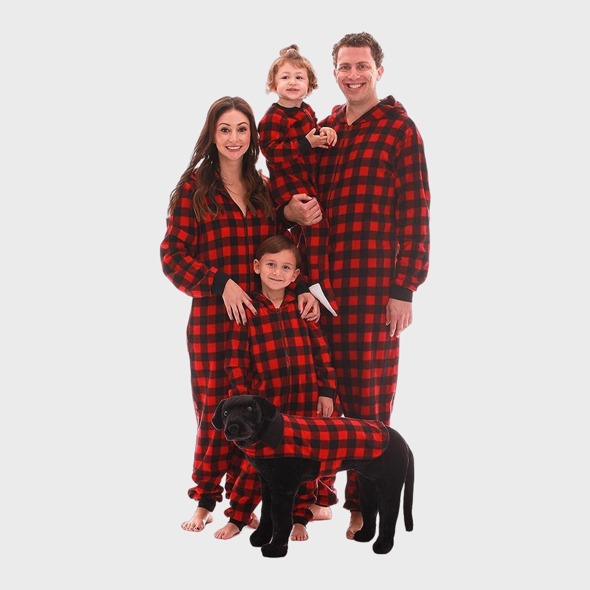 Family wearing matching red plaid pajamas, standing together with a black dog dressed similarly, against a plain white background.
