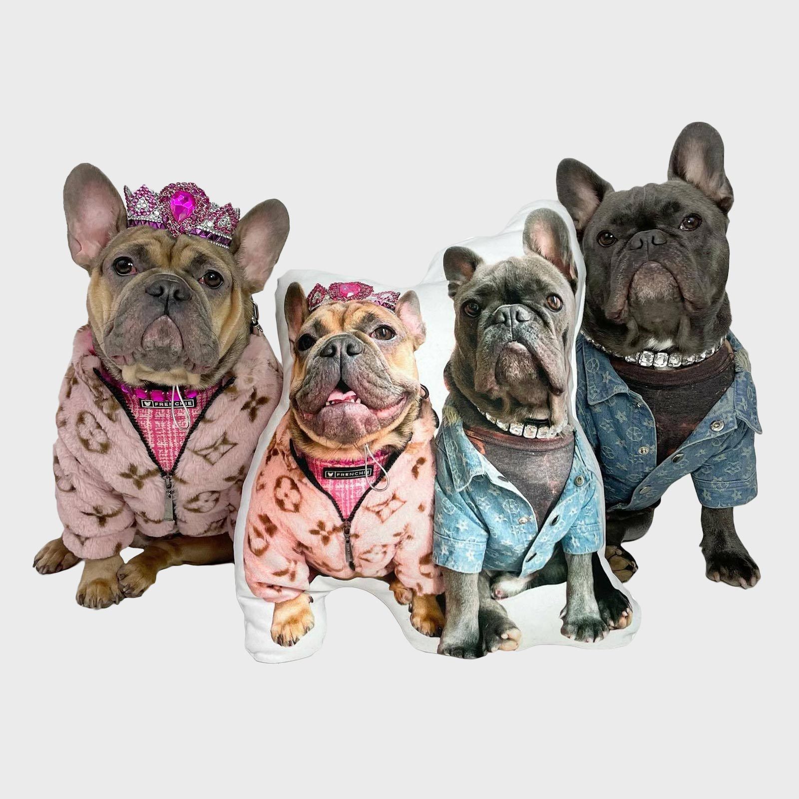 Two French bulldogs wearing jeweled tiaras and elaborate outfits sit beside matching dog-shaped pillows, against a plain background.
