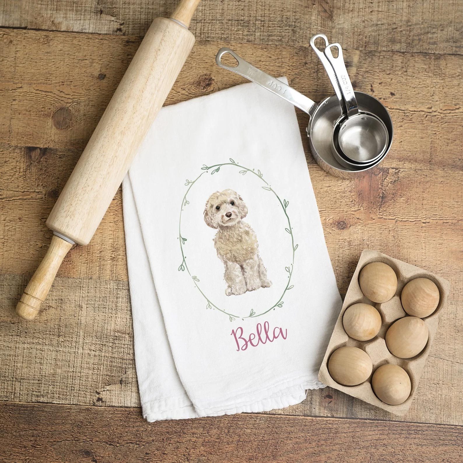 A rolling pin rests beside a dish towel with a dog illustration and the word "Bella," accompanied by measuring cups and wooden eggs on a wooden surface.