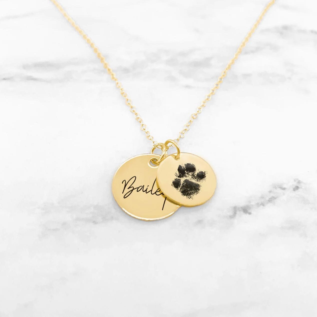 Gold necklace displays two round pendants; one inscribed with "Bailey," the other features a black paw print, set against a marble background.