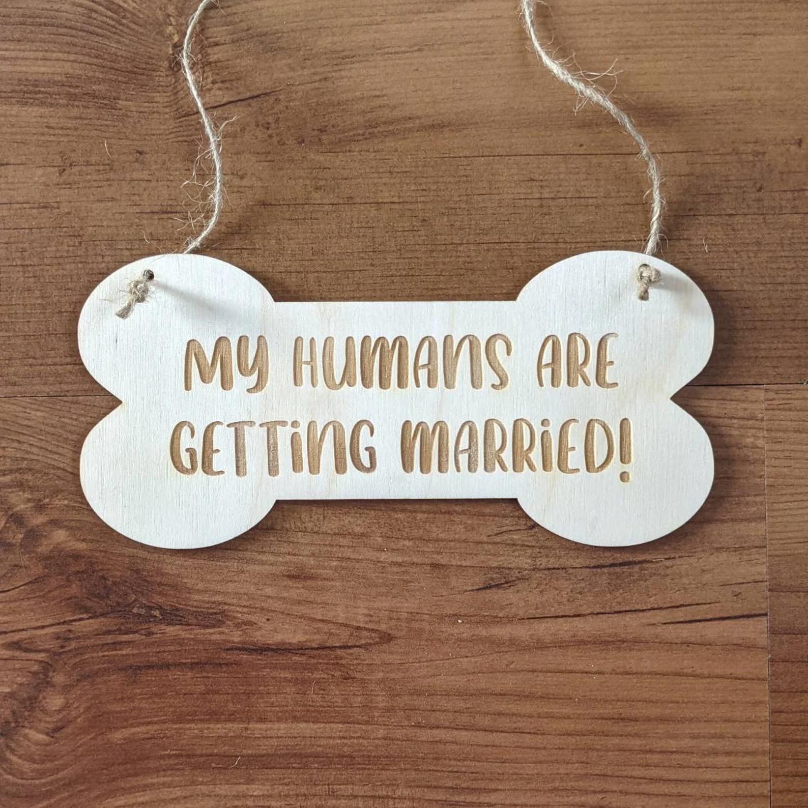Bone-shaped sign reads "MY HUMANS ARE GETTING MARRIED!" hanging on a wooden background.