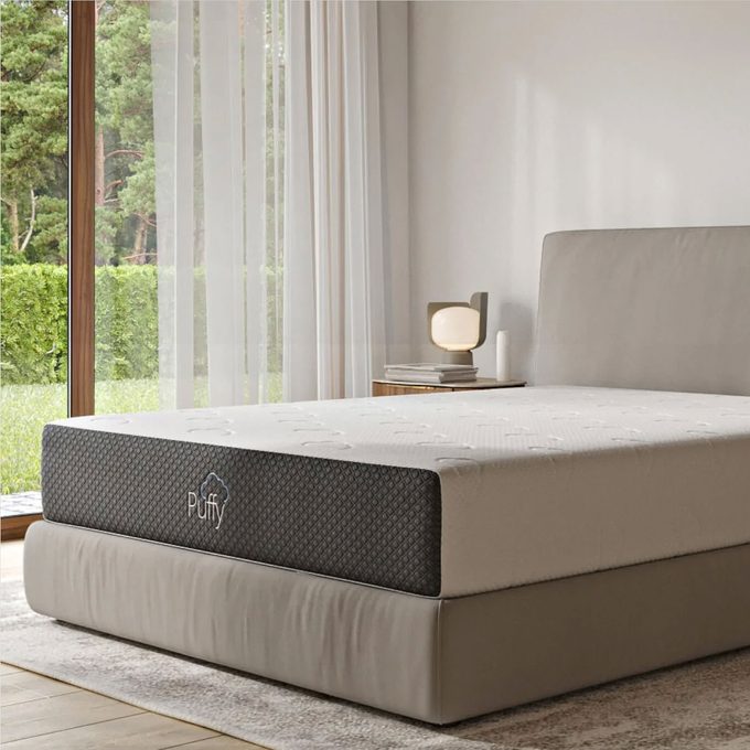 Mattress rests on bed, surrounded by soft lighting, a side table with books, and large windows overlooking greenery. Label reads 