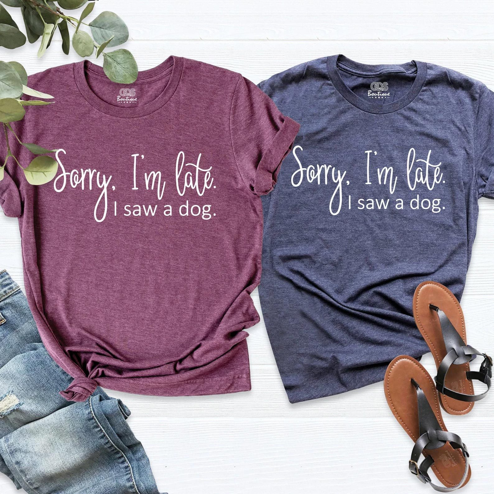 Two t-shirts lie on a white surface, each with text: "Sorry, I'm late. I saw a dog." Shorts and sandals nearby.