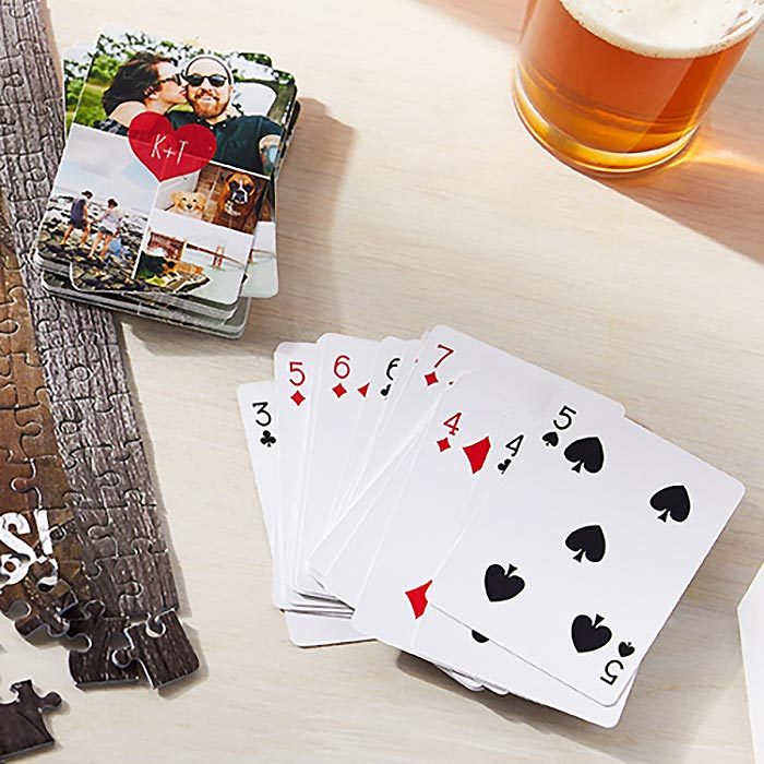 Playing cards lie fanned on a table near a puzzle, beside a stack of photo cards and a glass of beer.