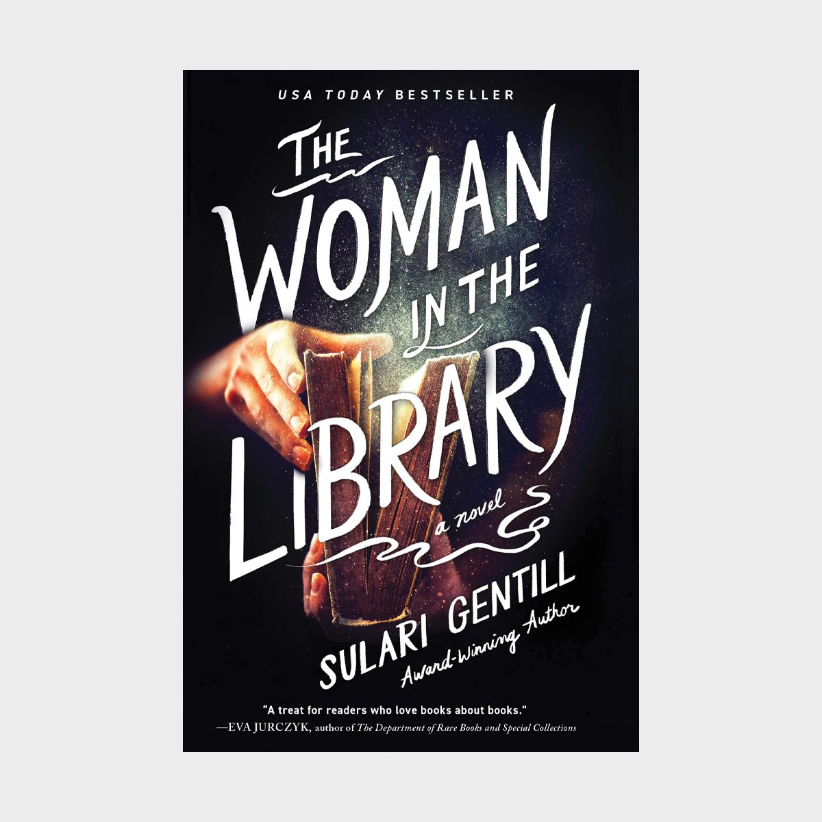 Hand holding book, showcasing title "The Woman in the Library" by Sulari Gentill. Dark background emphasizes book cover text.